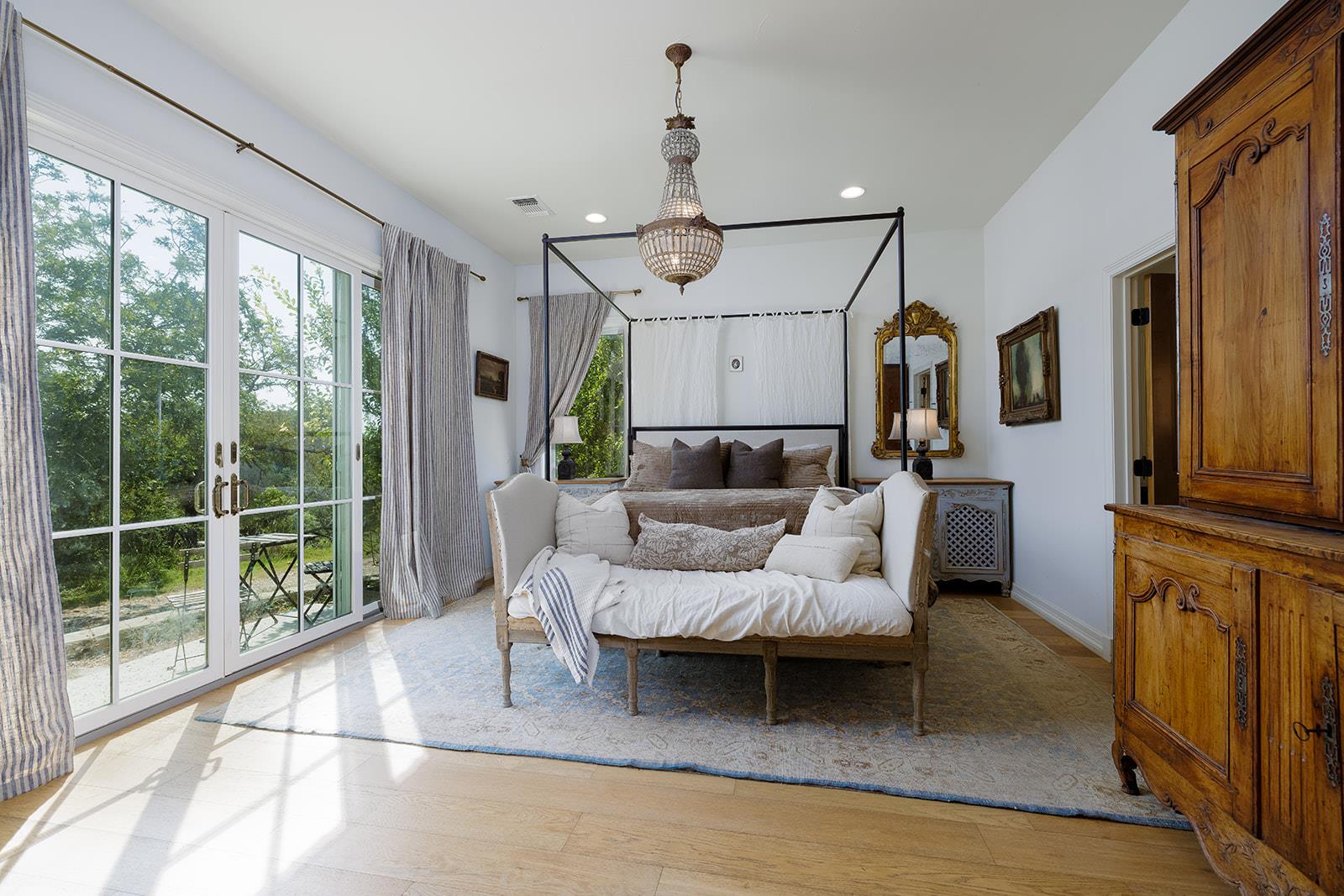 Bedroom with a four-poster bed, ornate wooden armoire, and tall glass doors overlooking greenery.