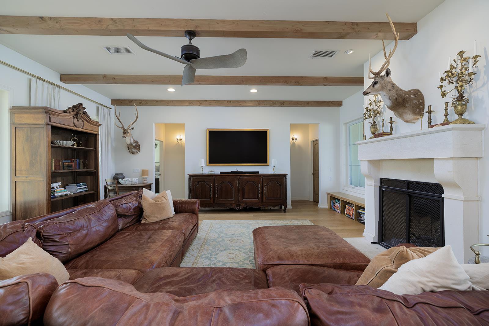 Living room with exposed wood beams, a brown leather sectional, white fireplace, and mounted deer heads.