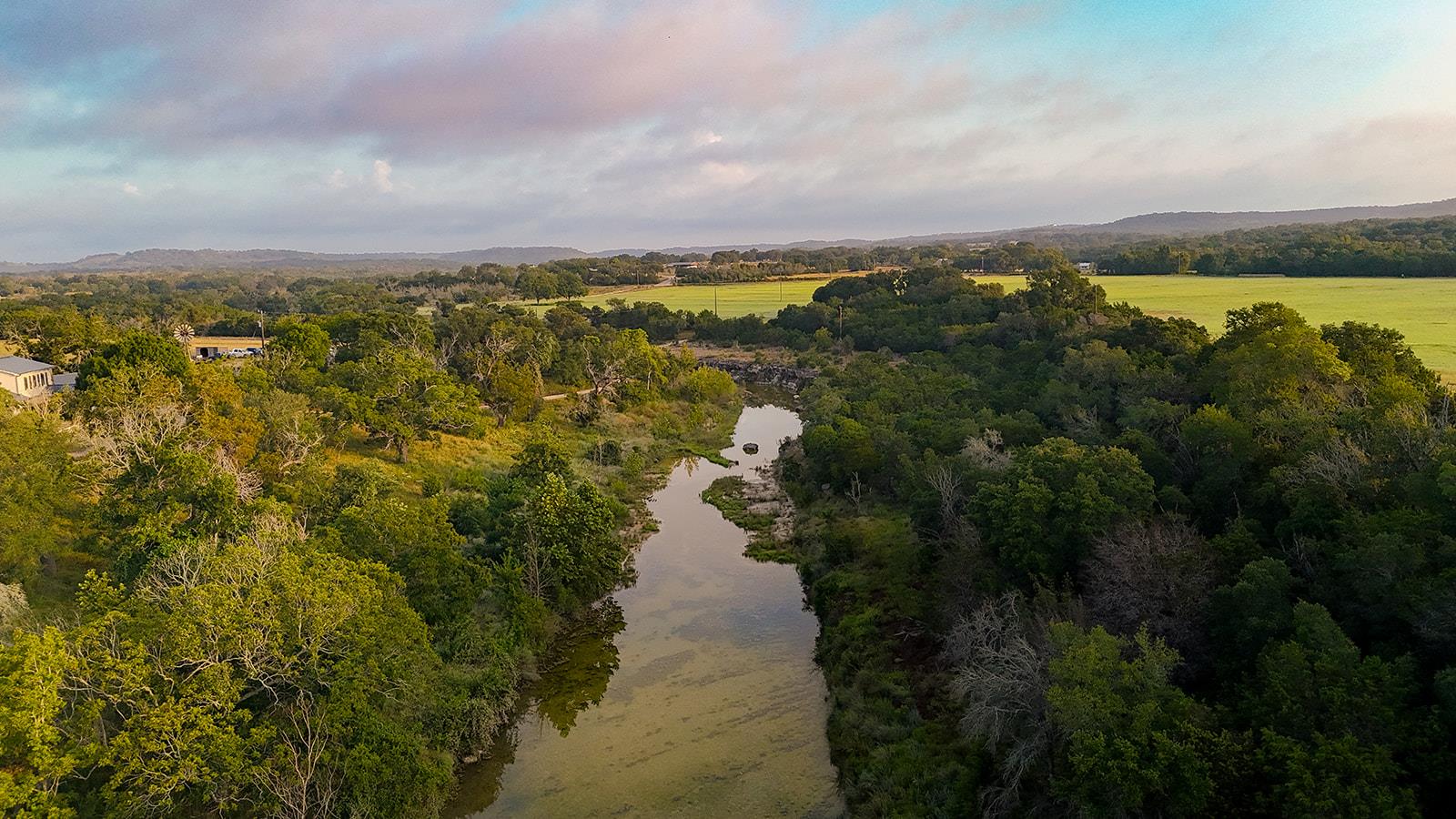 Aerial view of a tree-lined creek winding through wooded land toward open fields.