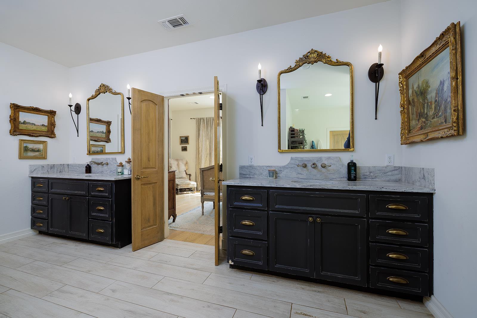 Bathroom with twin black vanities, pale stone counters, and ornate gold-framed mirrors.