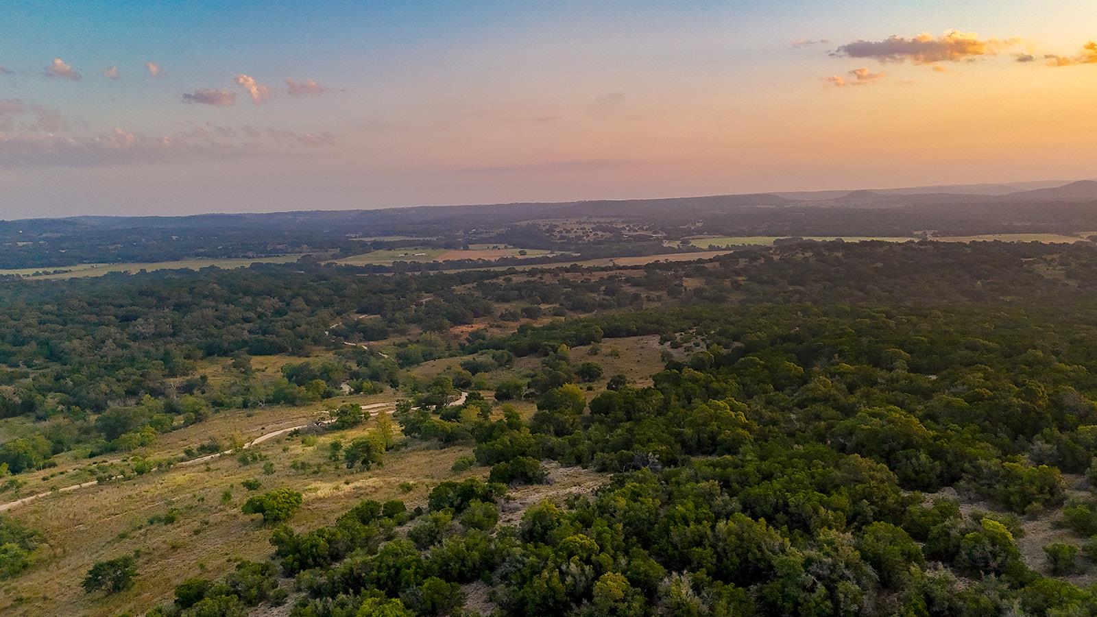Aerial view of rolling wooded acreage and open grassland beneath a pastel sunset sky.
