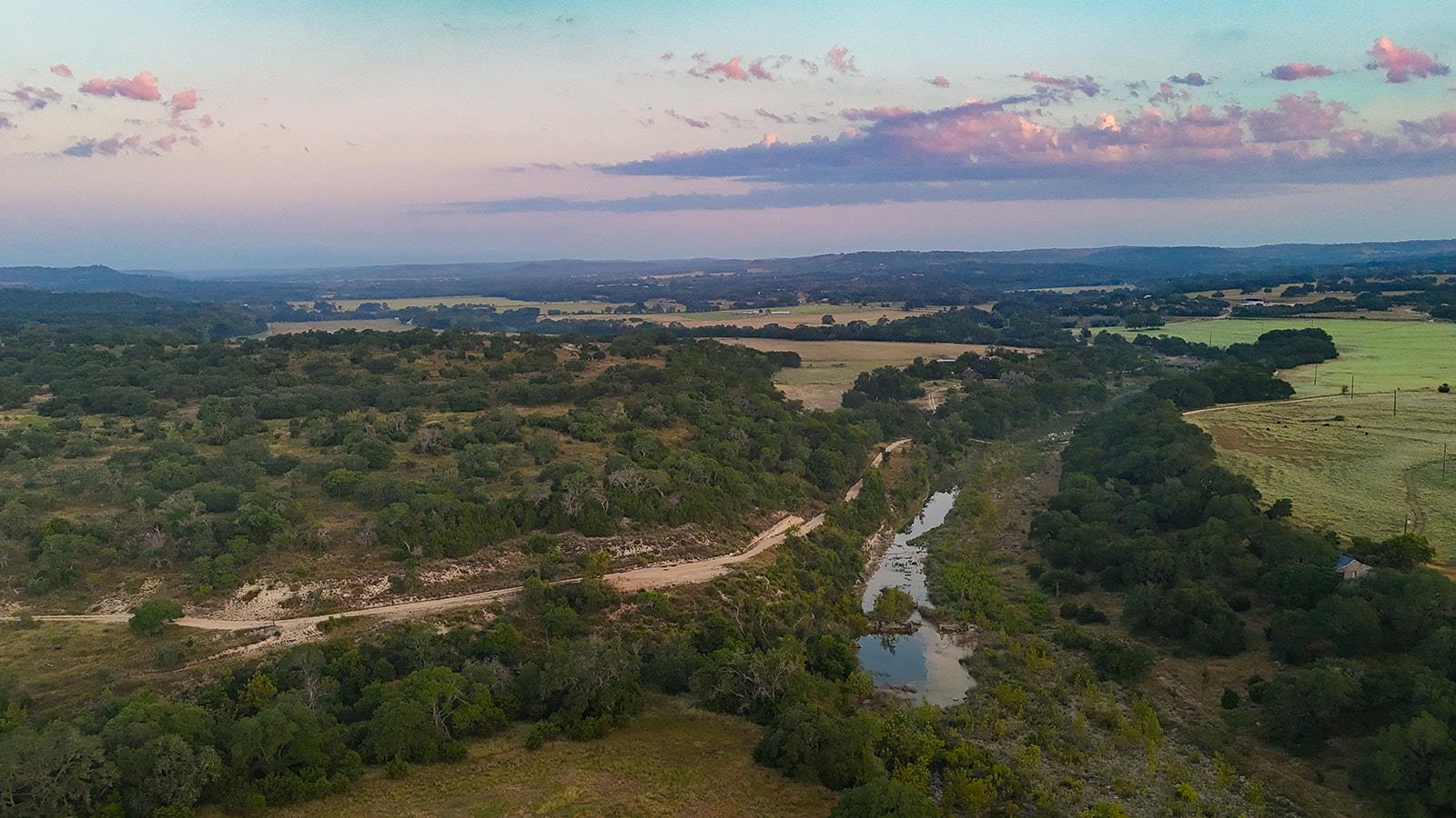 Aerial view of wooded ranch acreage, open pastures, a winding dirt road, and a creek at sunset.