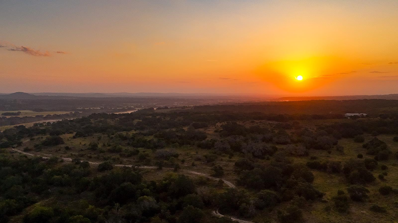 Aerial view of wooded ranch land and winding dirt tracks beneath a vivid sunset.
