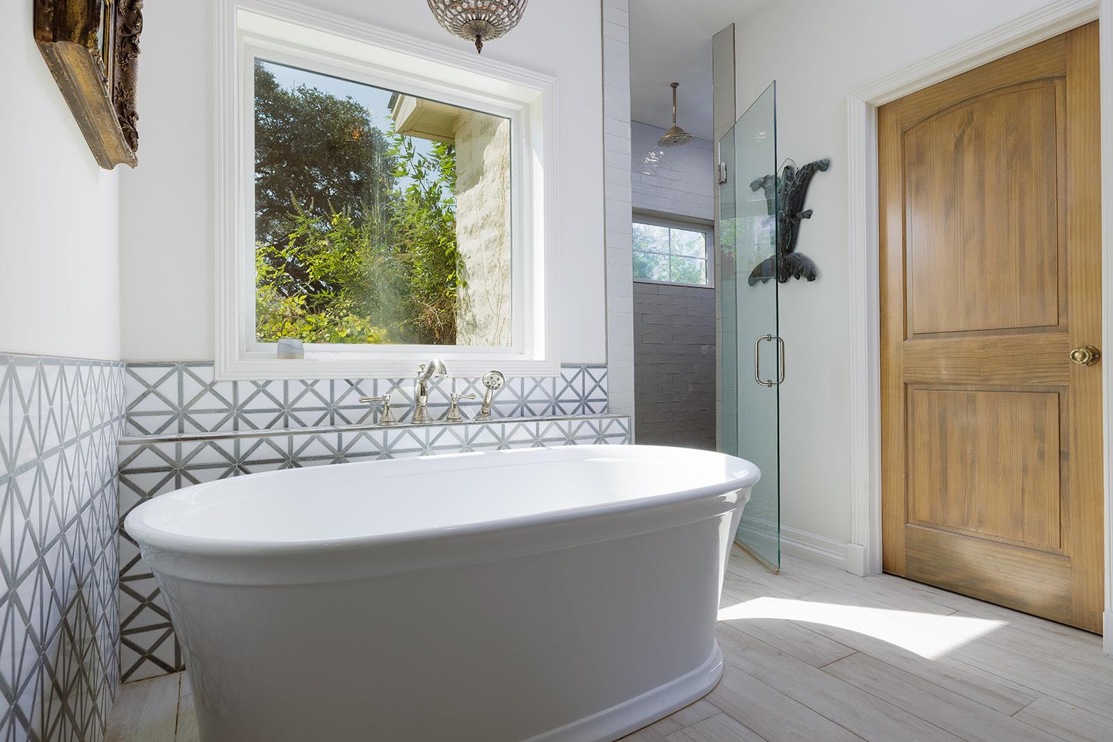 Bathroom with a freestanding tub, geometric tile, glass shower, and large window overlooking greenery.