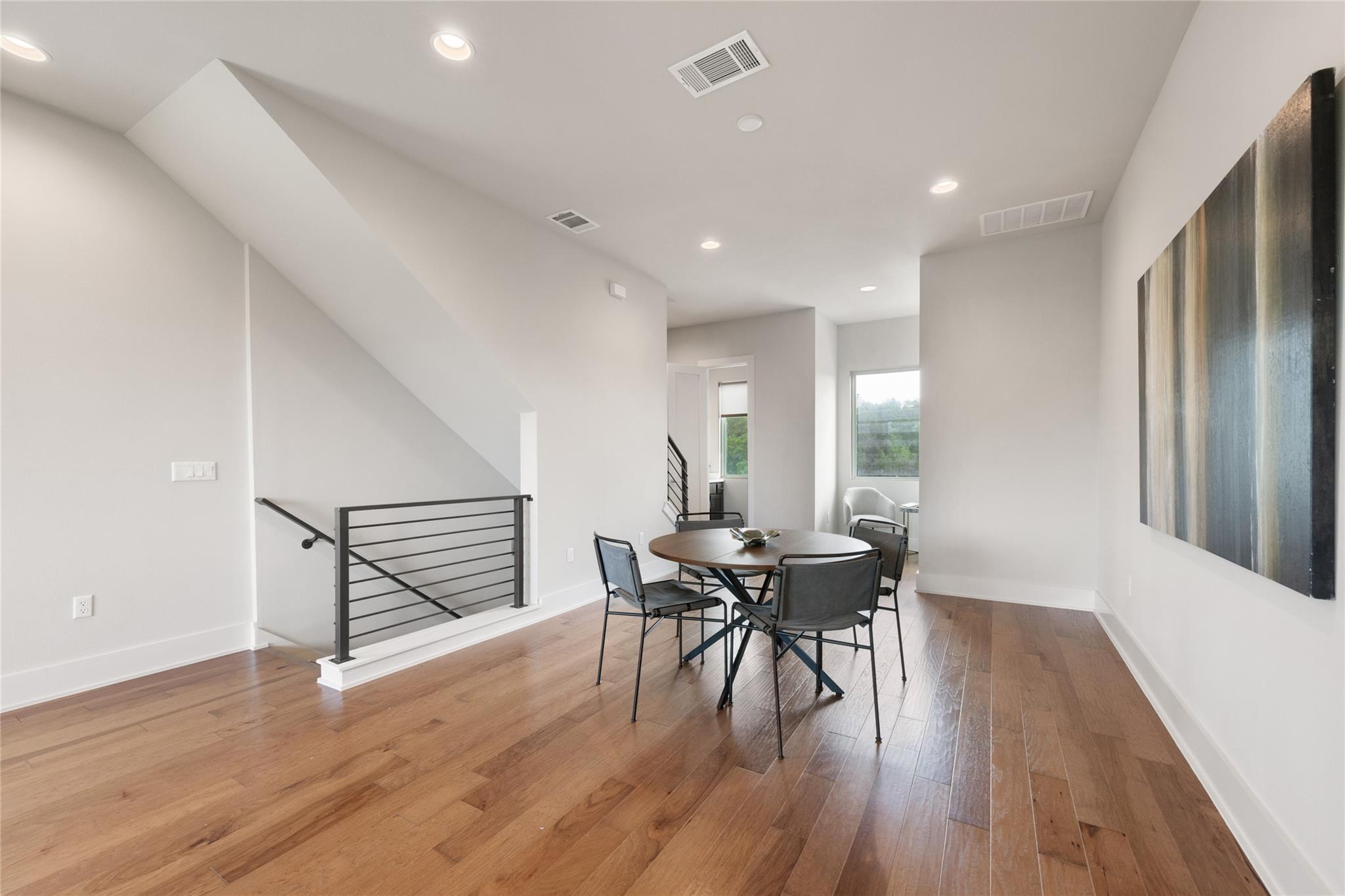 Dining area with a round table, wood floors, black stair railing, and large windows.