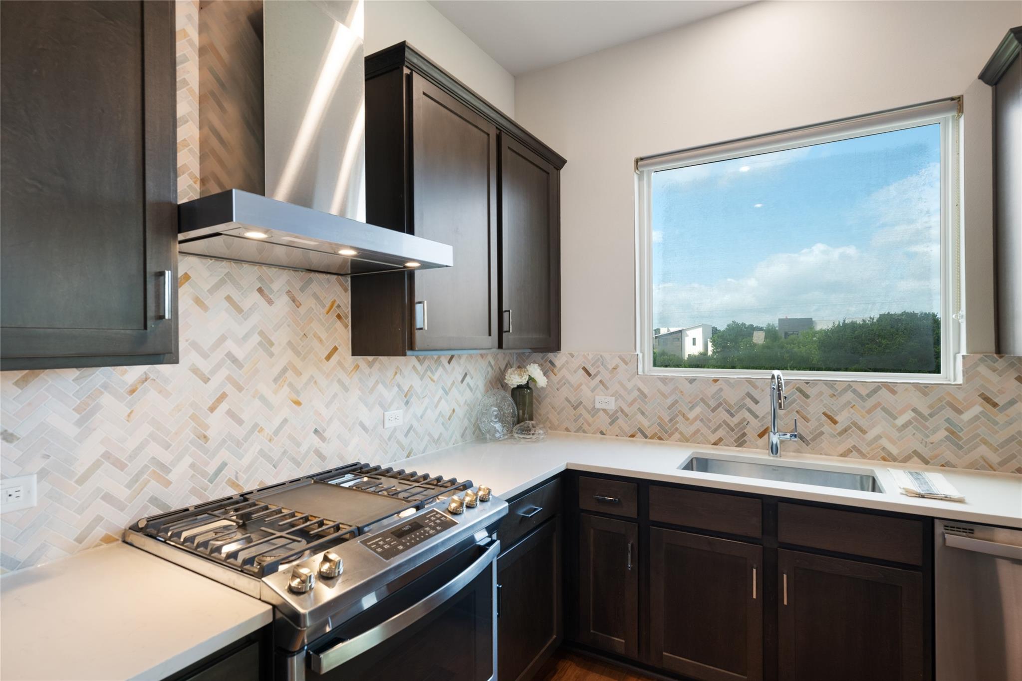 Modern kitchen with dark cabinets, geometric tile backsplash, stainless gas range, and a window above the sink.