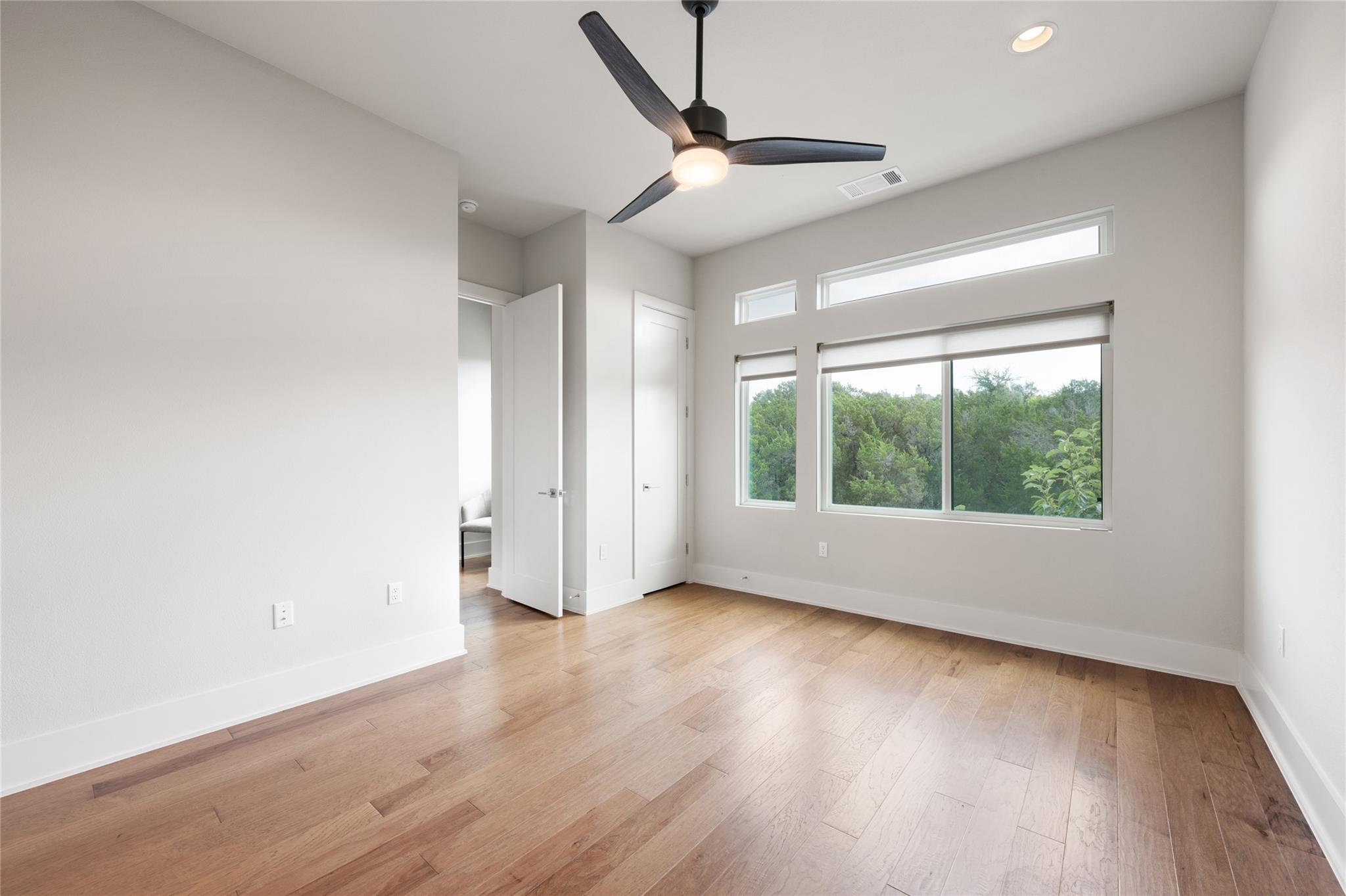 Bright empty room with wood floors, a ceiling fan, and wide windows overlooking greenery.