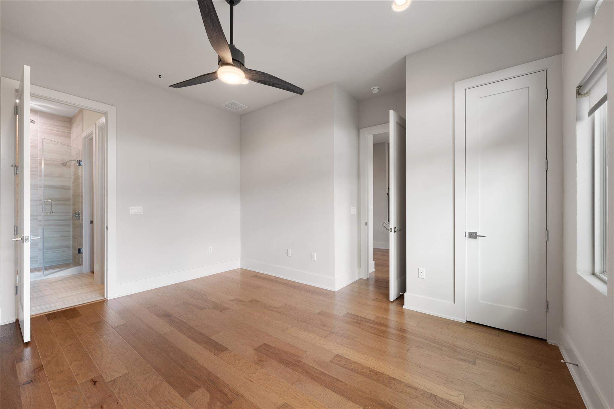 Empty bedroom with hardwood floors, a dark ceiling fan, and an open doorway to a tiled bathroom.