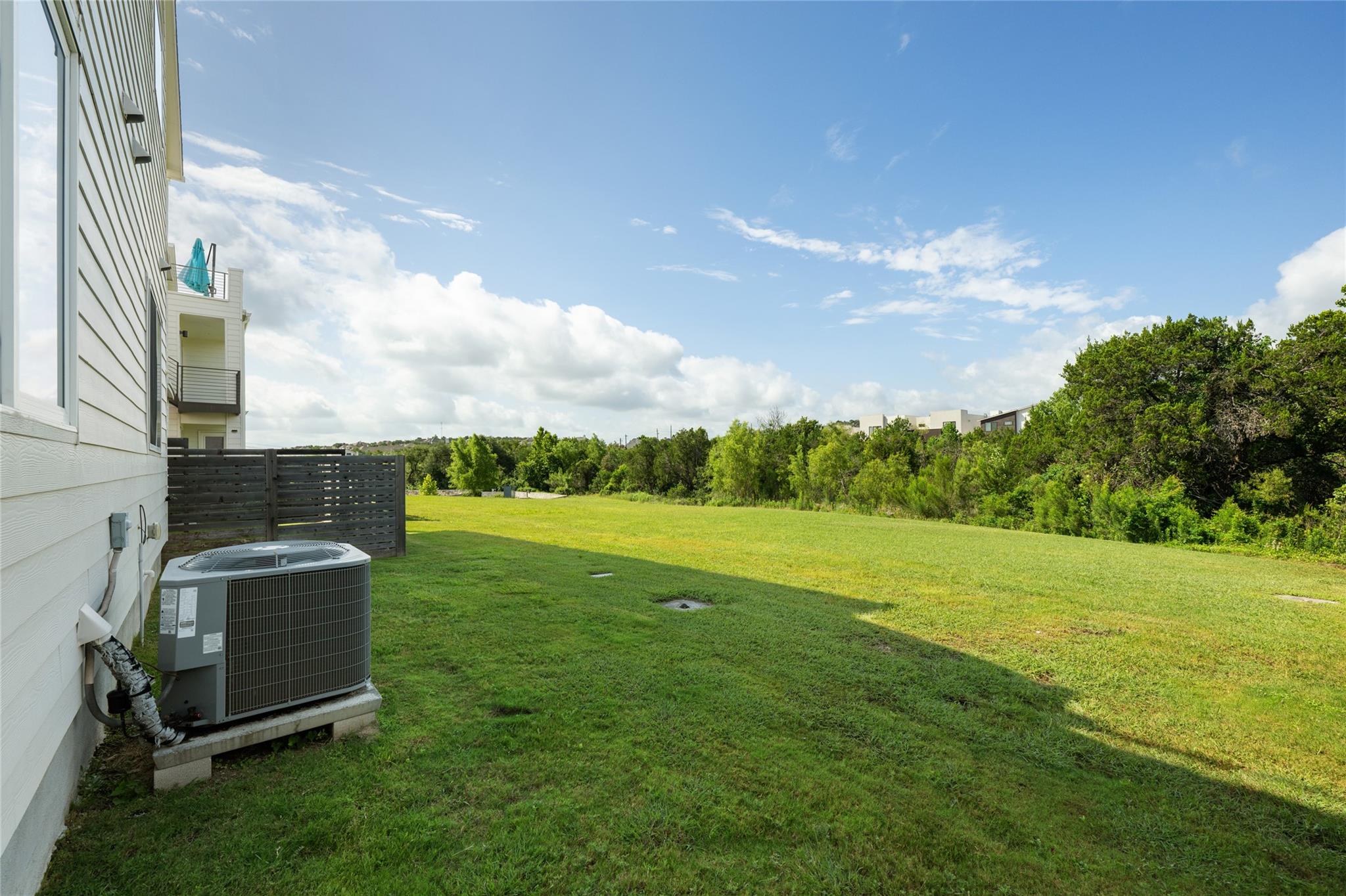 Grassy side yard beside a light-sided home, with a privacy fence and trees along the far edge.