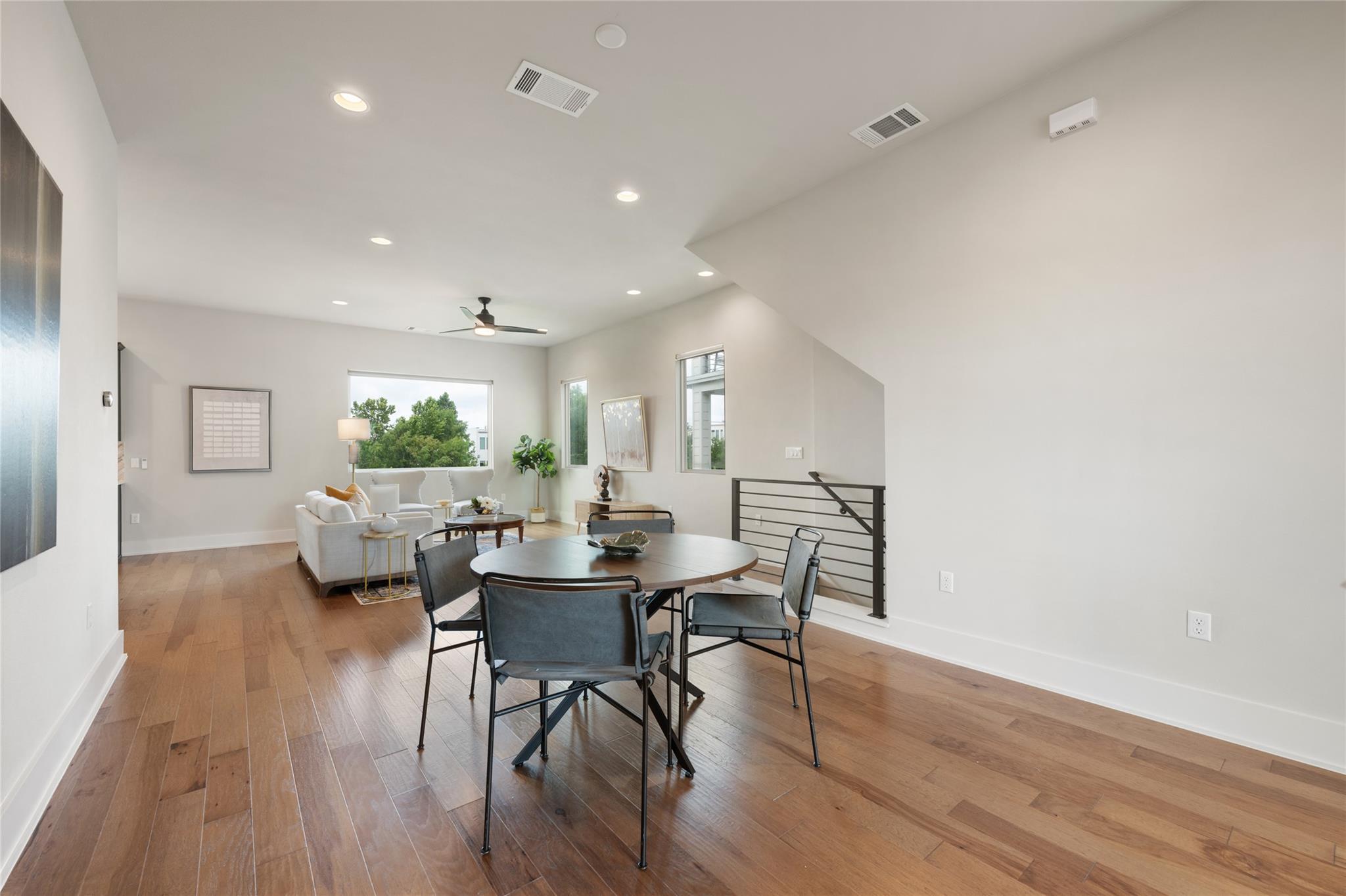 Open dining and living area with a round table, warm wood floors, white sofa, and black stair railing.