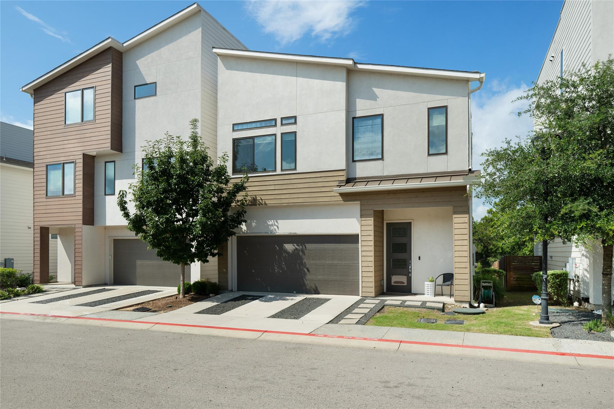 Modern multi-story home with white stucco, tan siding, two garages, and a small front tree.