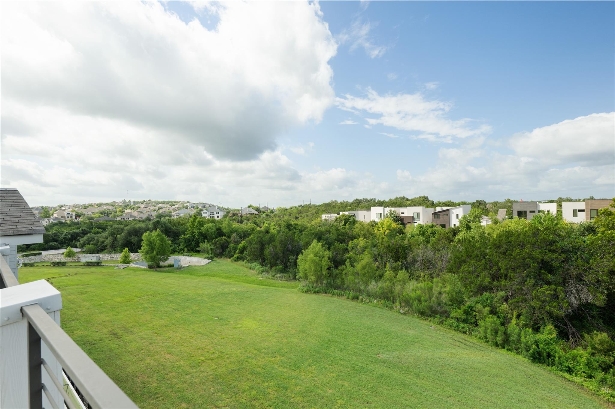 Balcony view over a broad grassy lawn and wooded greenbelt toward neighboring homes.