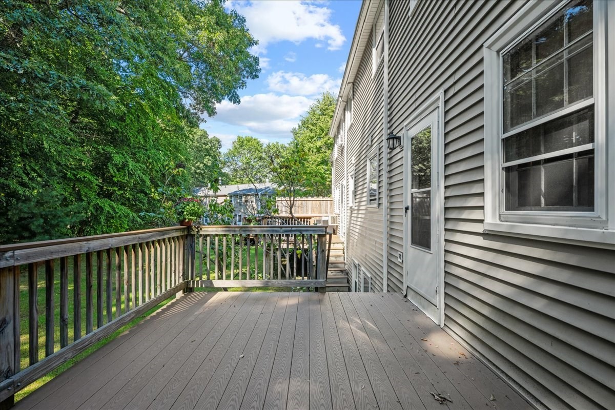 Elevated deck beside gray-sided home, with a wooden railing and leafy yard beyond.