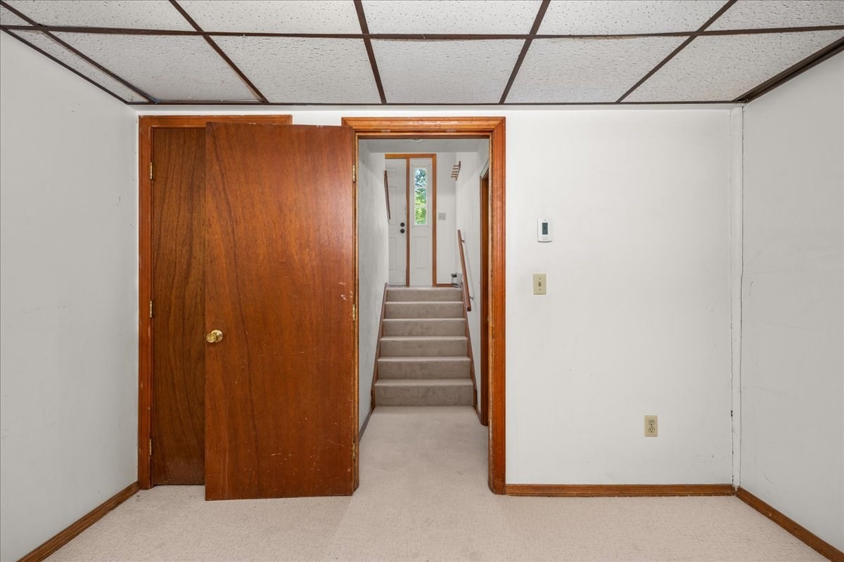 Empty carpeted room with an open wooden door revealing stairs and a white door beyond.