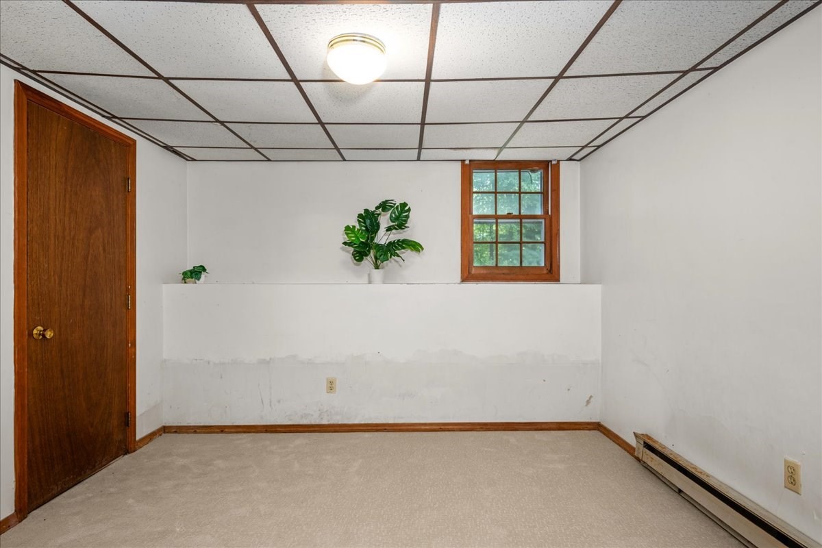 Empty carpeted room with a grid ceiling, wooden door, small window, and baseboard heater.