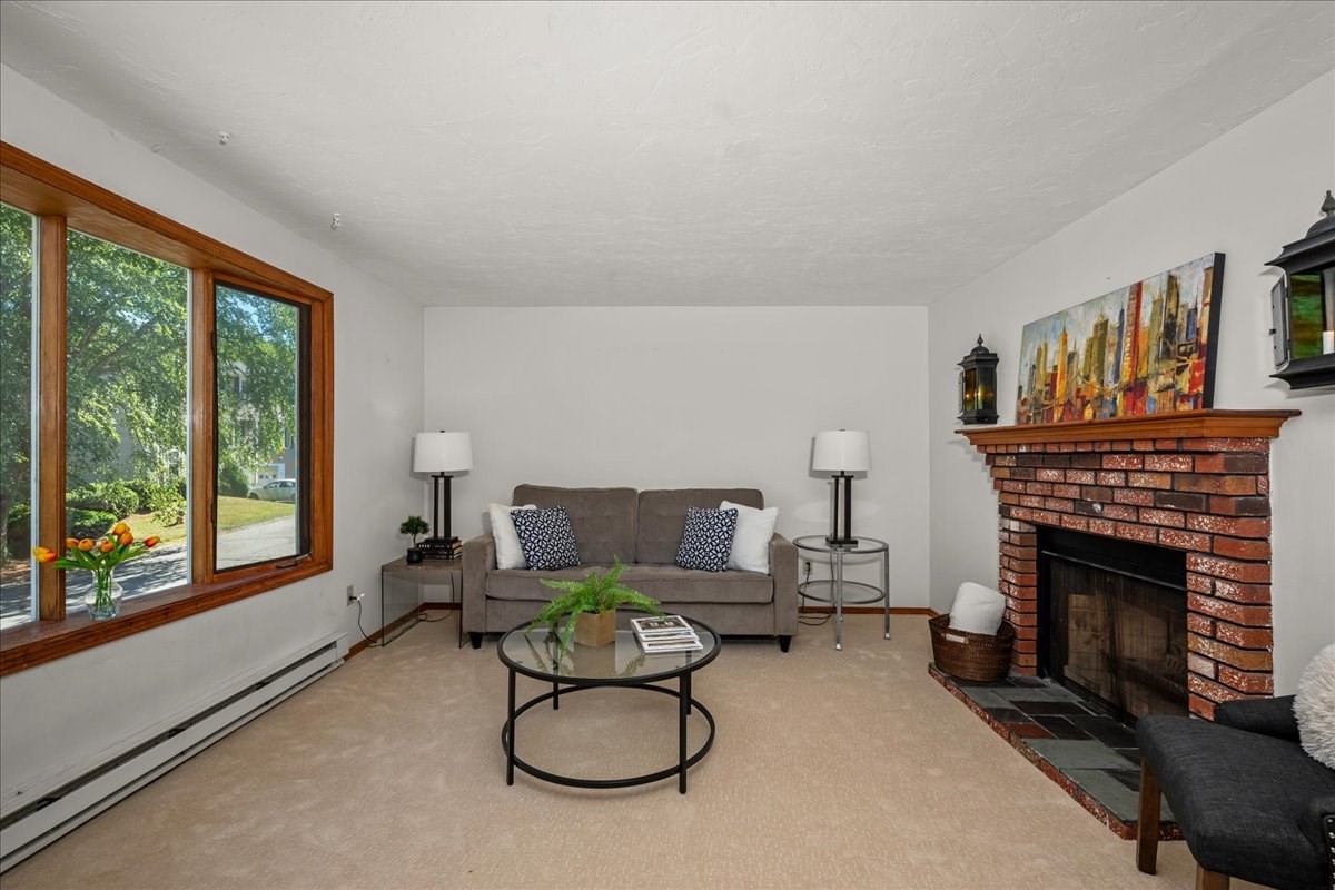 Living room with a brick fireplace, gray sofa, and large wood-framed window overlooking a leafy yard.