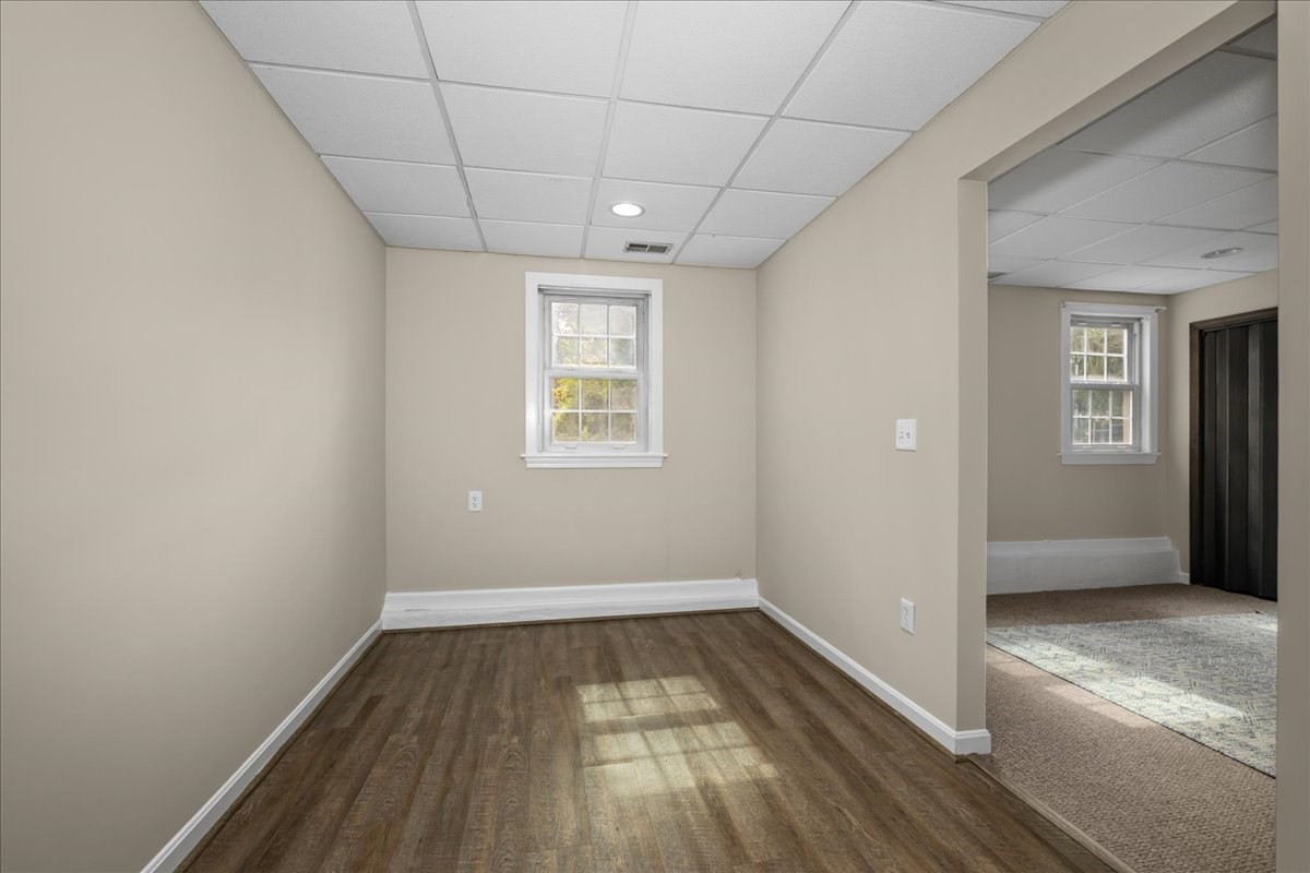 Empty room with dark wood-look floors, a suspended ceiling, and a wide opening to a carpeted room.