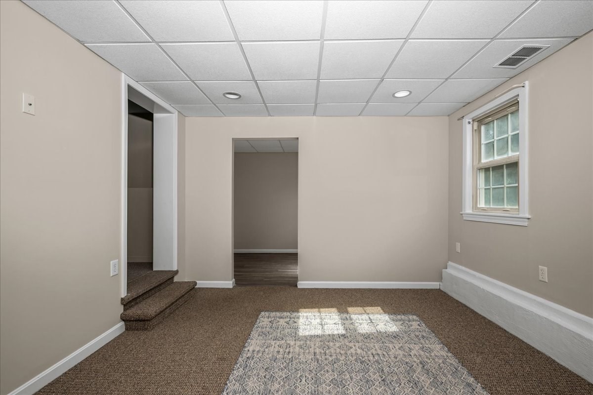 Empty carpeted room with a drop ceiling, window, patterned rug, and short steps to a raised area.