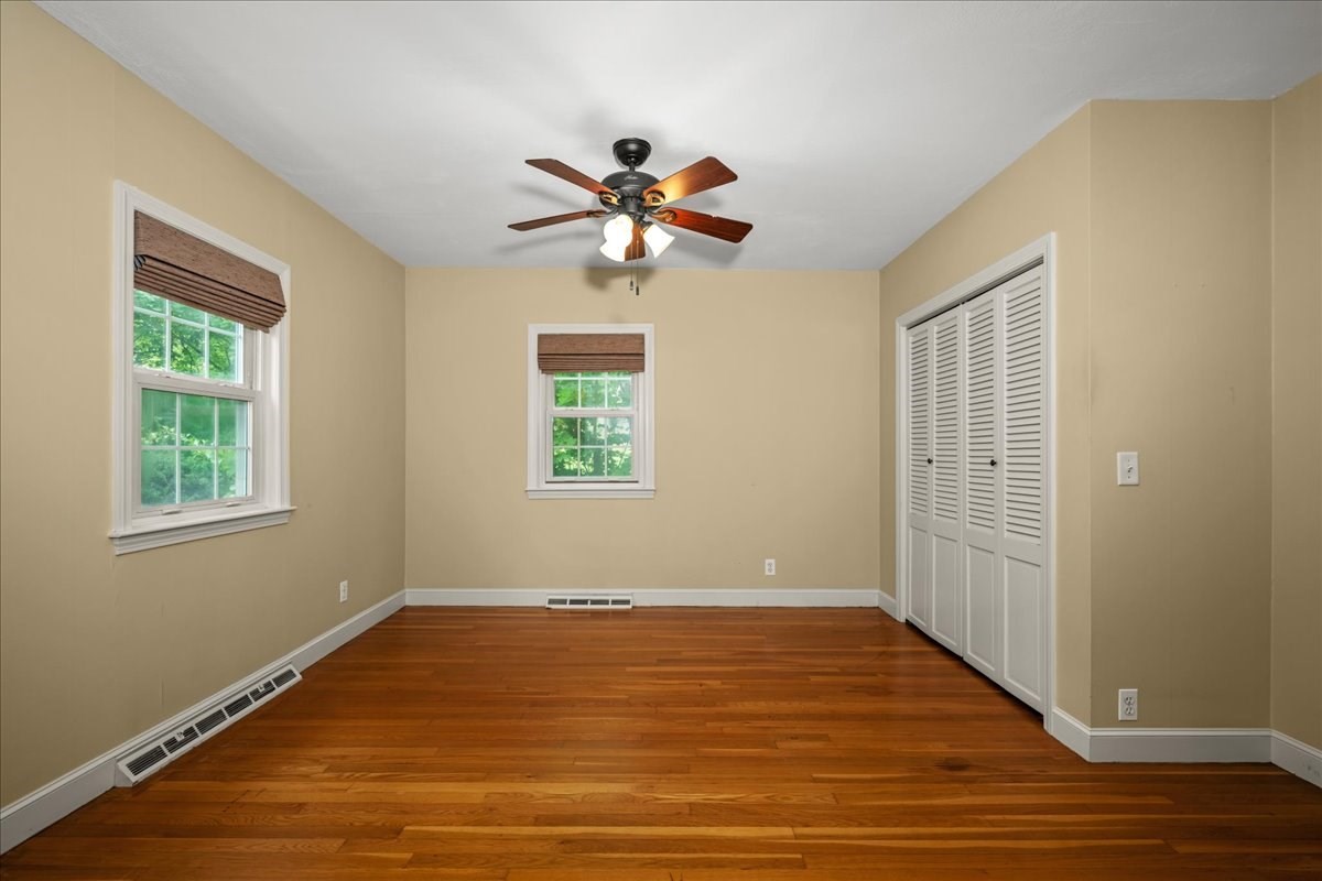 Empty bedroom with hardwood floors, two windows, a ceiling fan, and louvered closet doors.