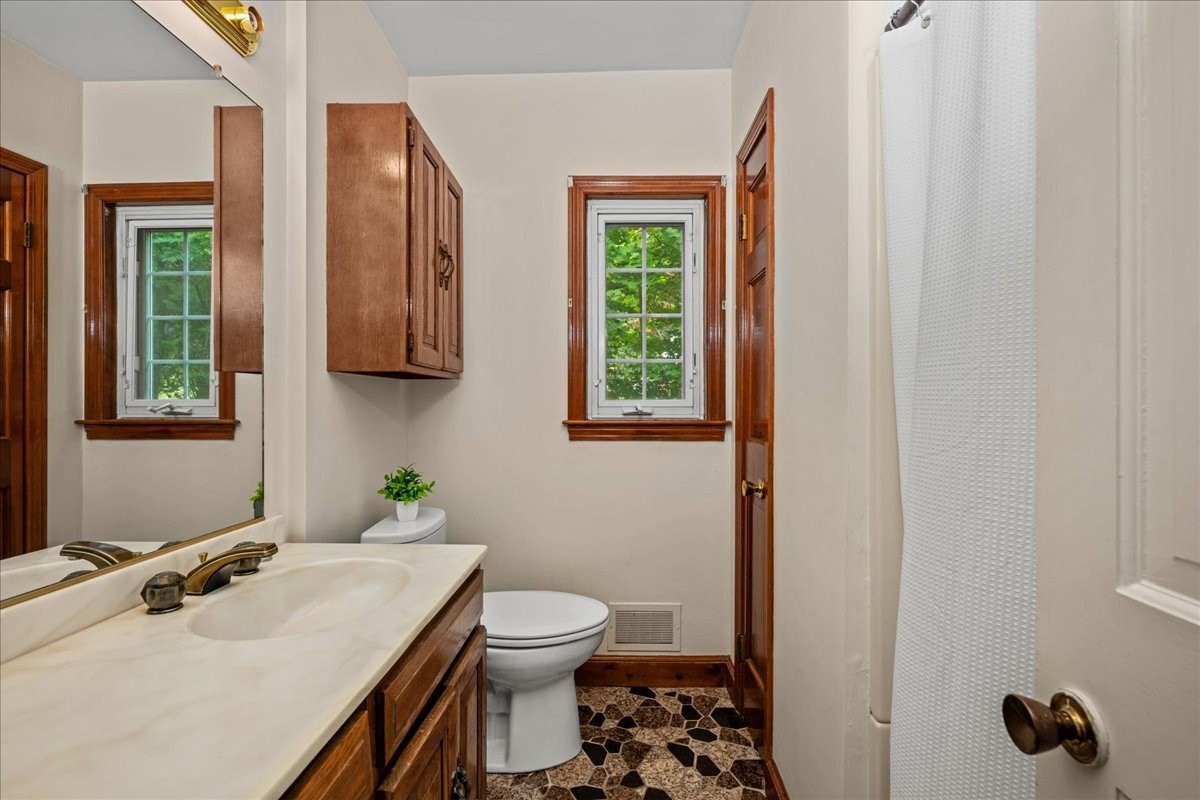 Bathroom with a long white vanity, dark wood cabinets, toilet, and windows overlooking greenery.