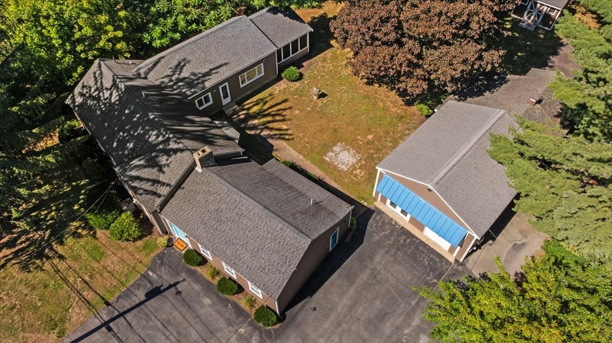 Aerial view of several buildings with gray roofs, a blue-sided outbuilding, and paved parking among trees.