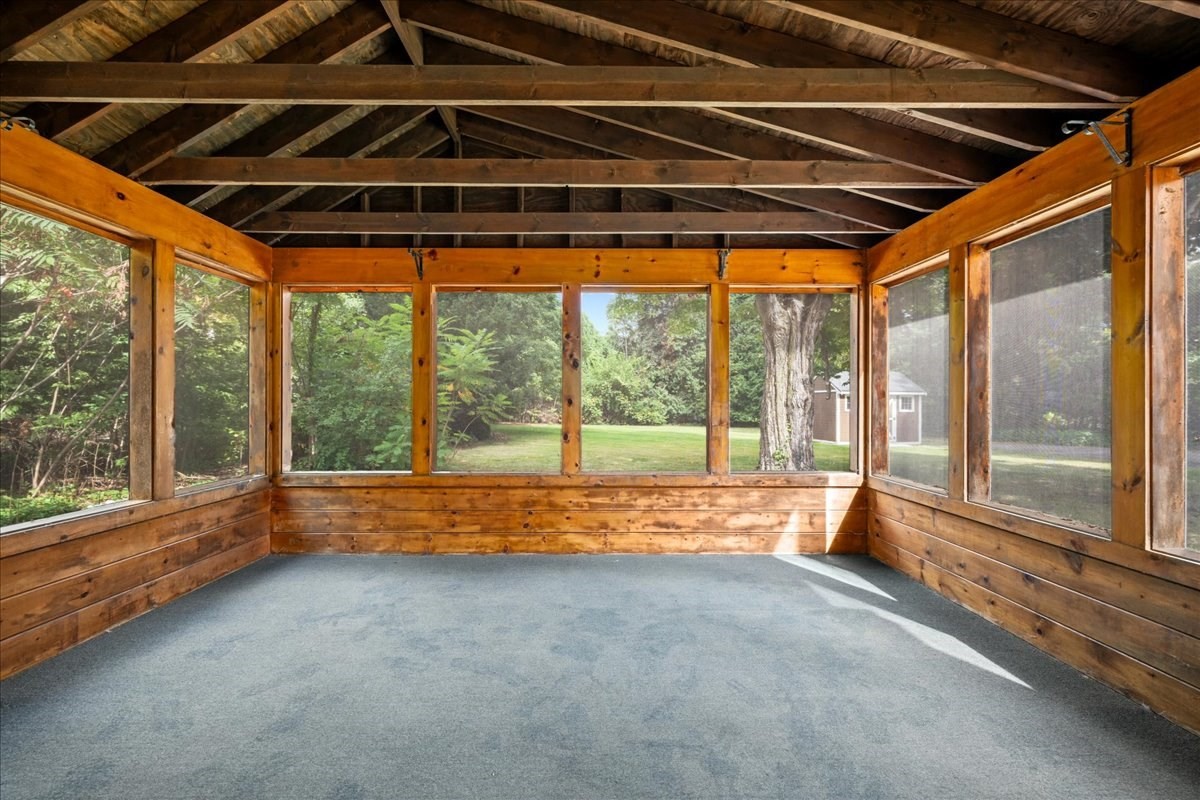 Enclosed sunroom with exposed timber beams, wall-to-wall windows, and blue-gray carpet.