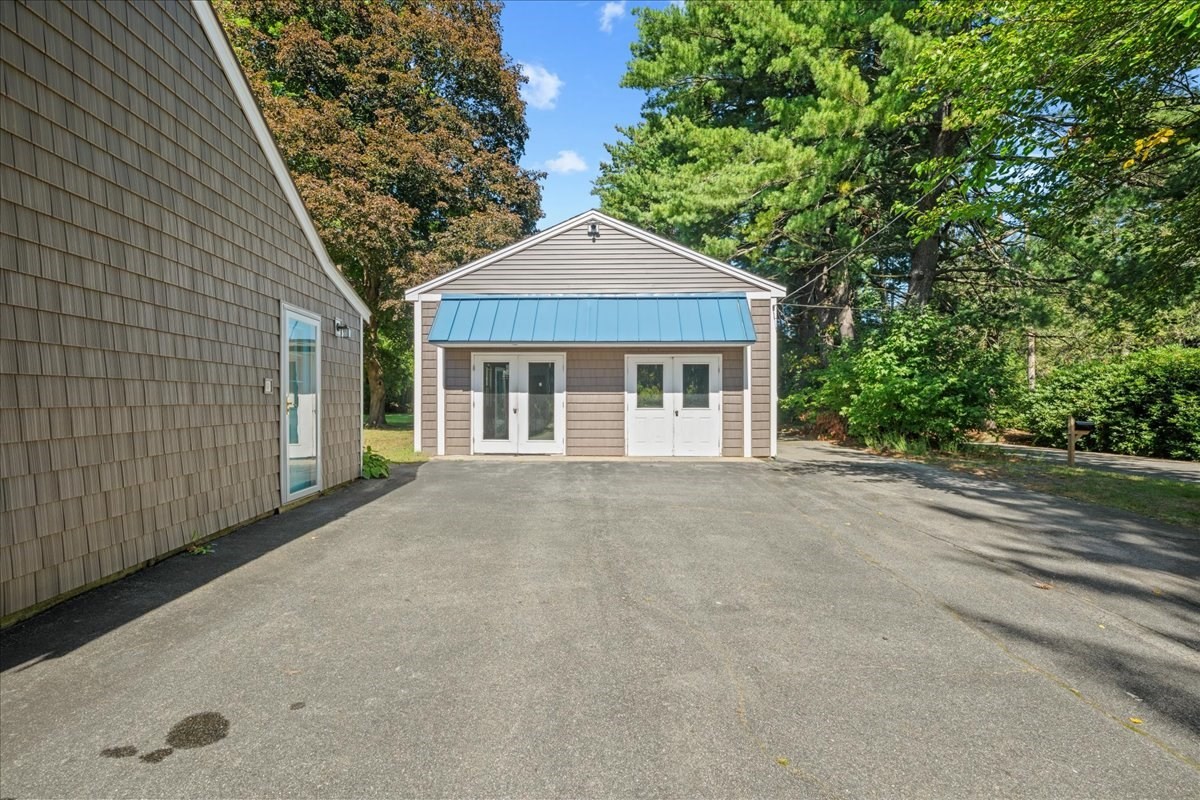 Small gray-shingled building with a blue awning and white doors, viewed across a paved driveway.