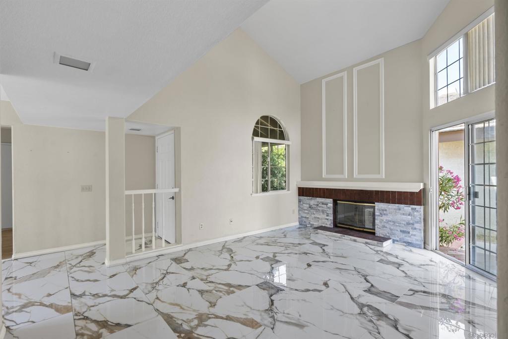 Vacant living room with vaulted ceiling, marble-pattern tile, stone fireplace, and sliding glass doors.