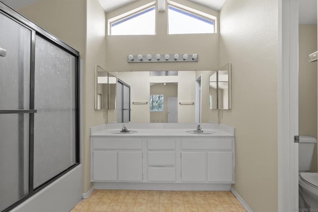 Bathroom with a double-sink vanity, mirrored wall, glass shower, and high windows.