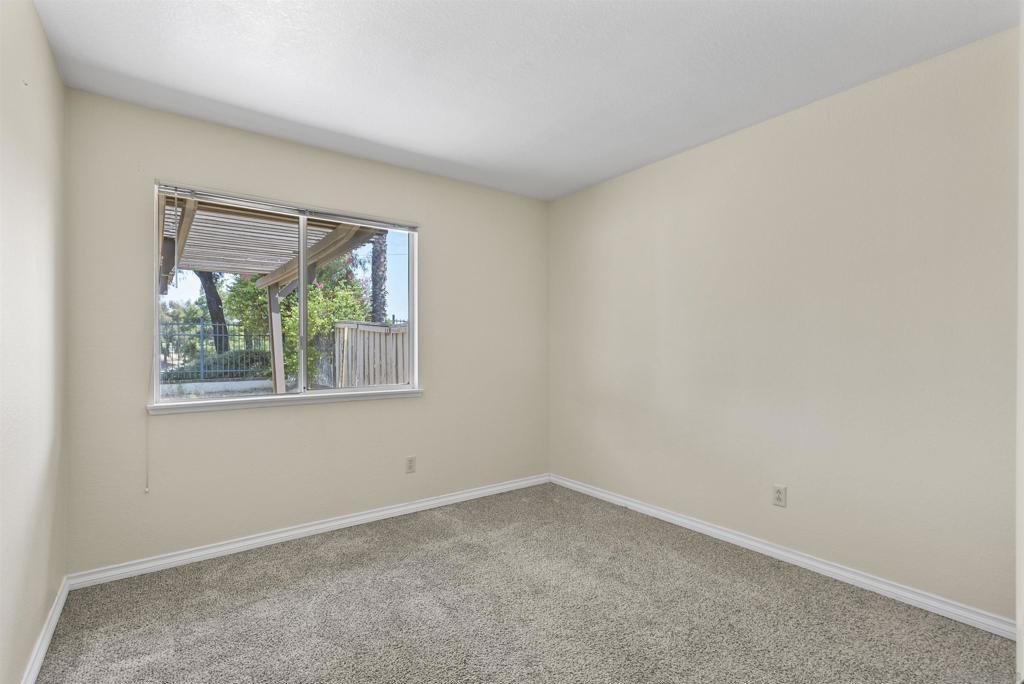 Empty bedroom with gray carpet and a window overlooking a fenced patio and trees.