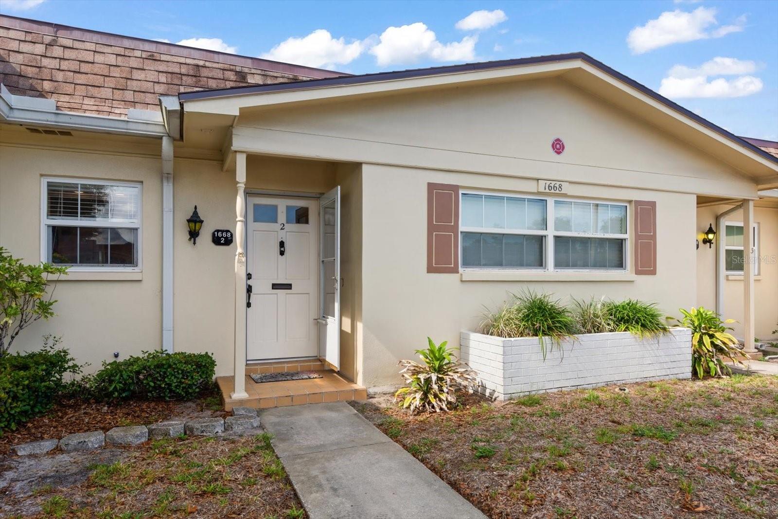 Front of a beige residential unit with brown shutters, a white entry door, and a raised planter.