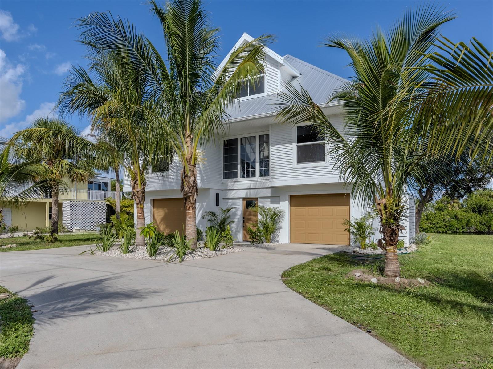 White coastal-style home with tan garage doors, framed by palm trees and a wide driveway.