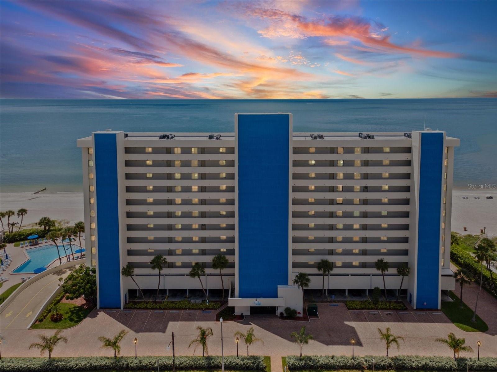 Blue-and-white beachfront condominium overlooking the Gulf, with a pool and colorful sunset sky.