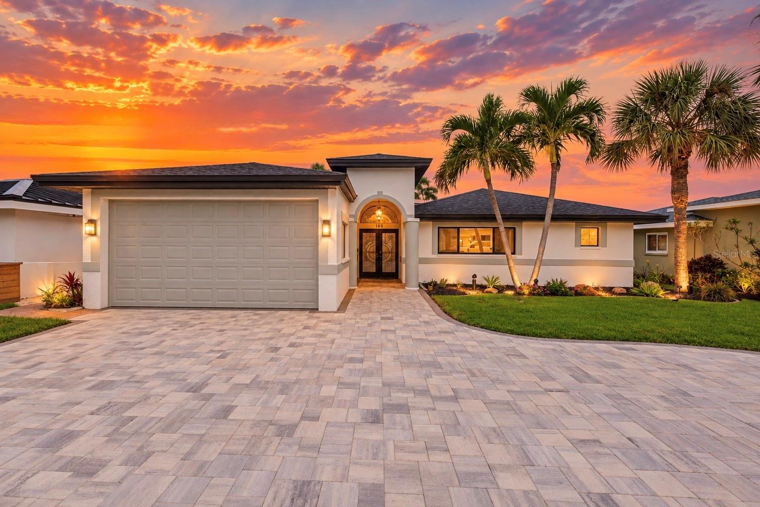 Single-story home with a wide paver driveway and palm trees beneath a vivid sunset.