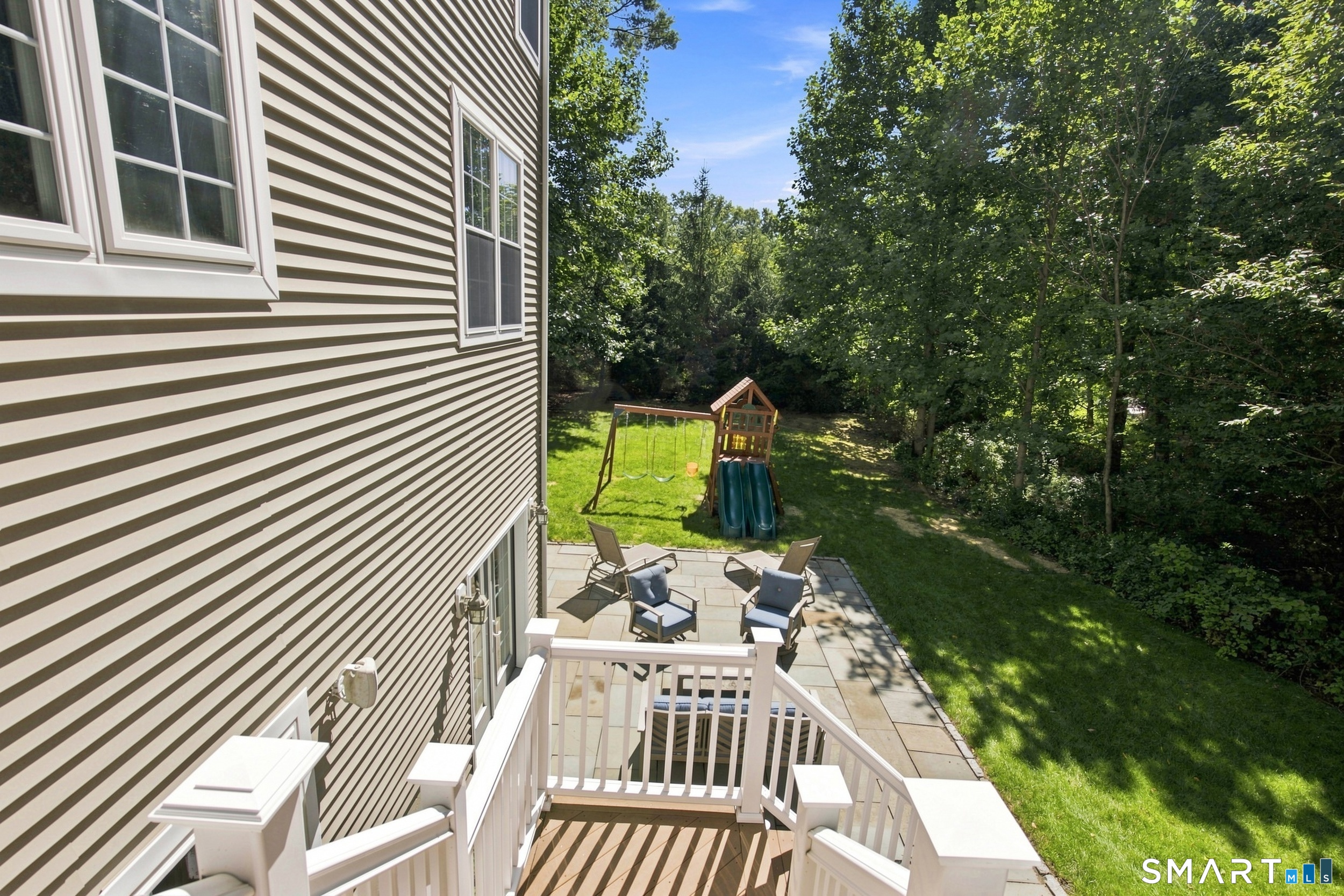 Raised deck overlooks a stone patio, grassy backyard, and children's playset bordered by mature trees.