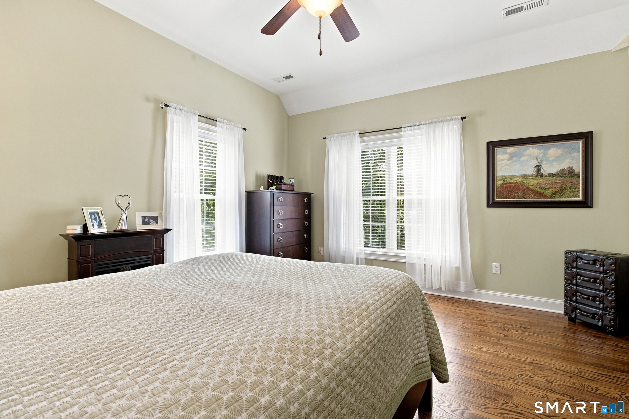 Bedroom with a large patterned bed, tall curtained windows, hardwood floors, and dark wood furniture.