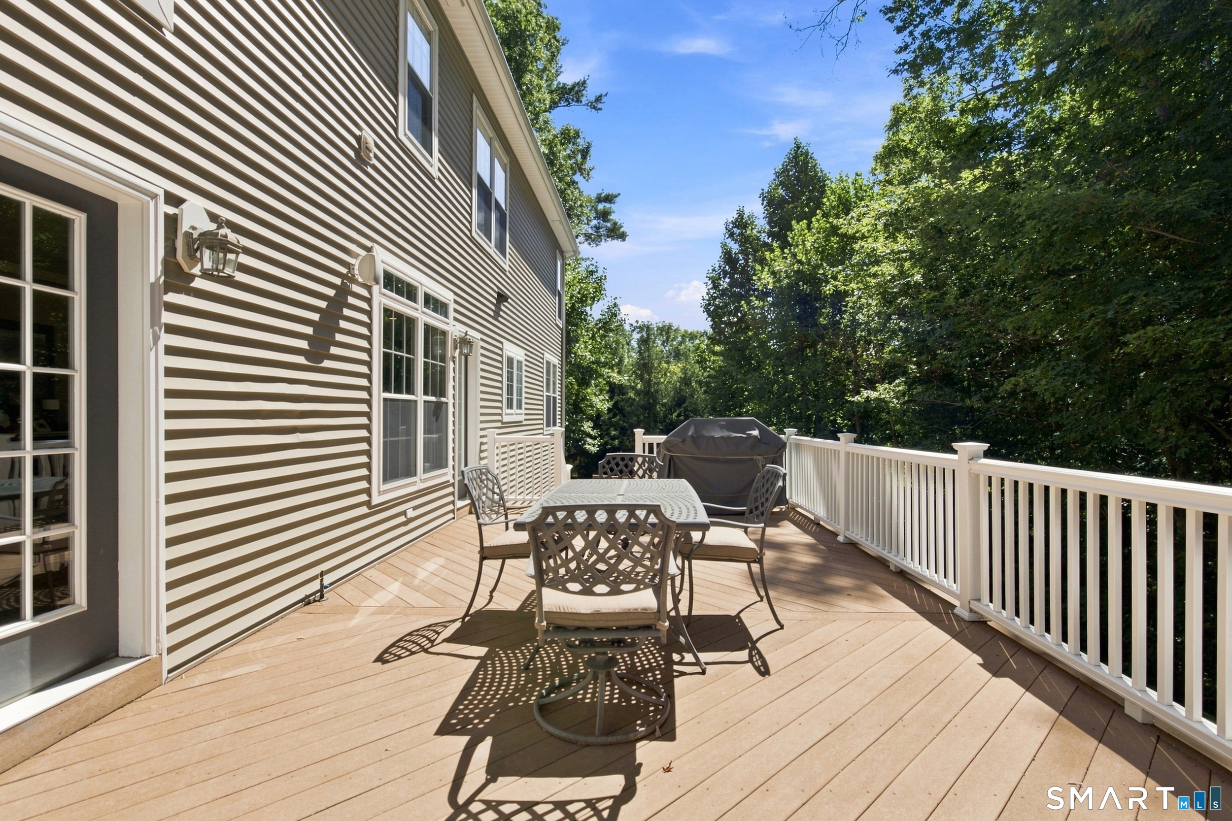 Raised deck with patio dining set and covered grill, bordered by white railing and leafy trees.