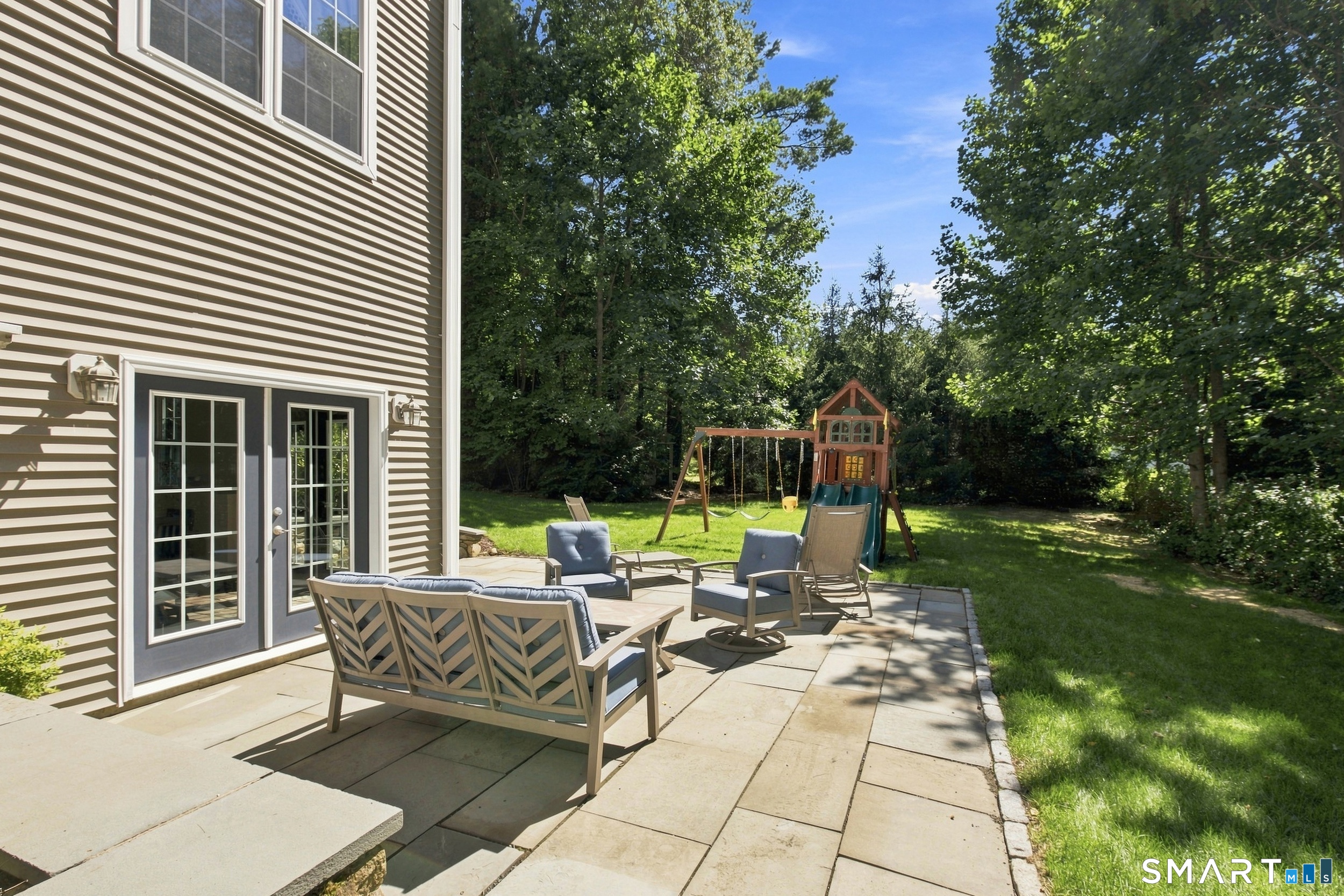 Backyard patio with outdoor seating and a children’s playset in a tree-lined lawn.