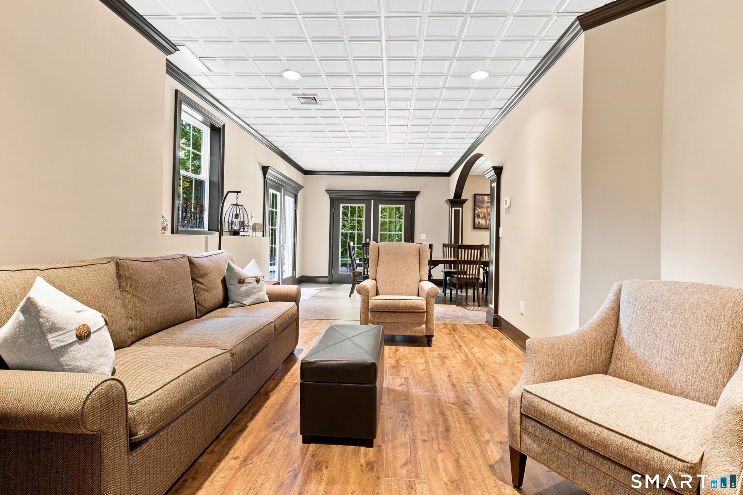 Furnished living room with a brown sofa, beige armchairs, coffered ceiling, and wood-look floors.