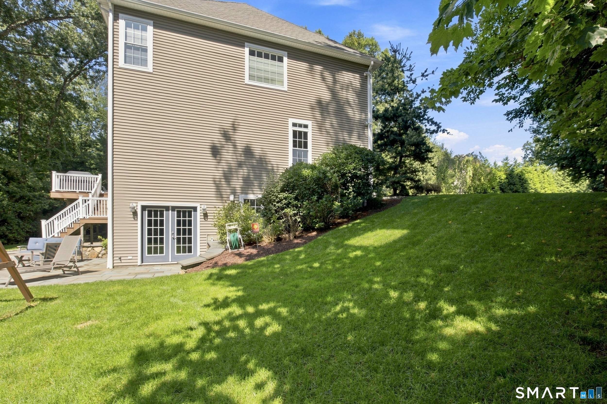 Rear exterior of a beige-sided house beside a sloping lawn, patio, and white-railed deck stairs.
