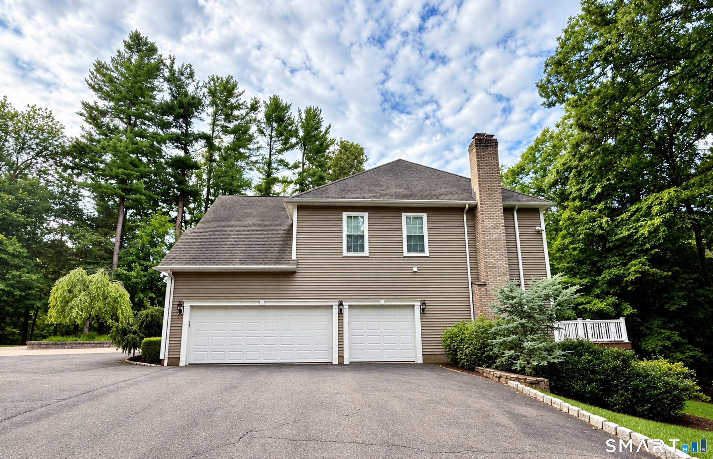 Taupe-sided house with a two-car garage and tall brick chimney, surrounded by mature trees.