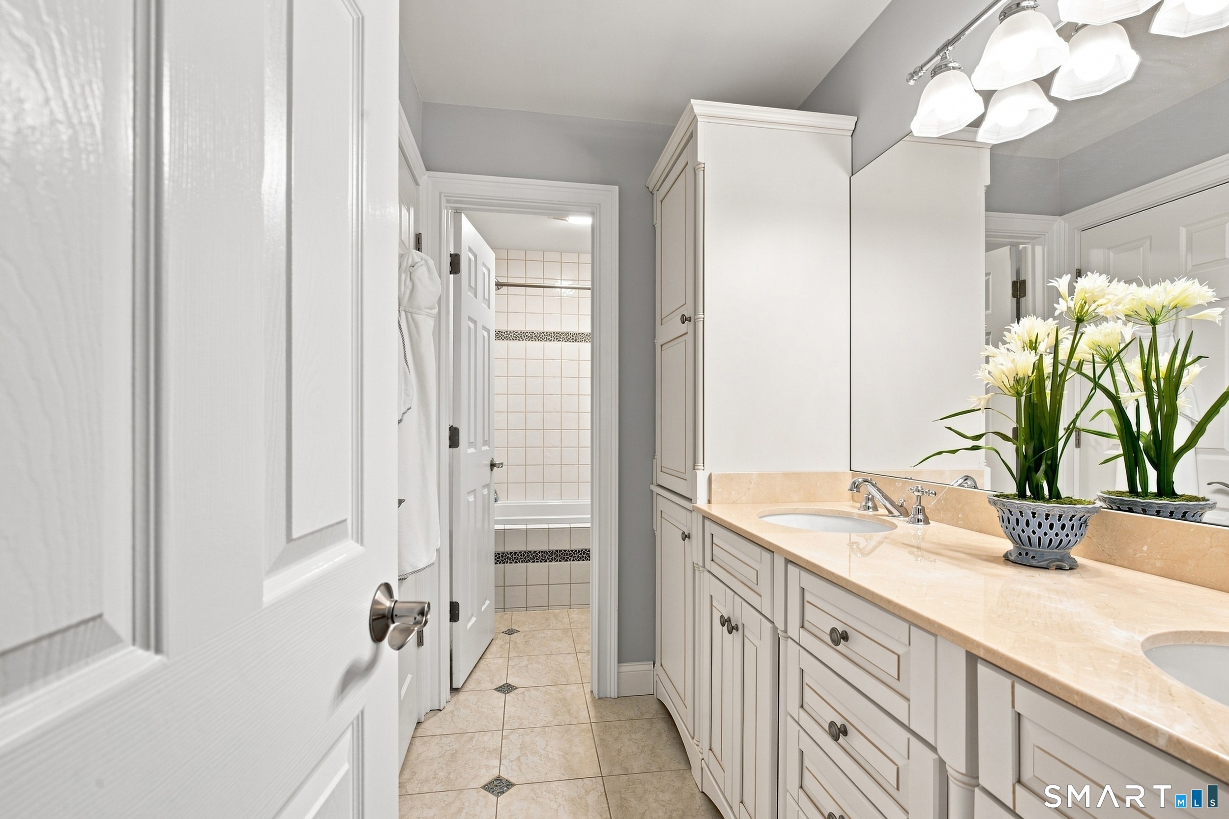 Bathroom vanity with white cabinetry, cream stone counter, and flowers beside a doorway to the tub.