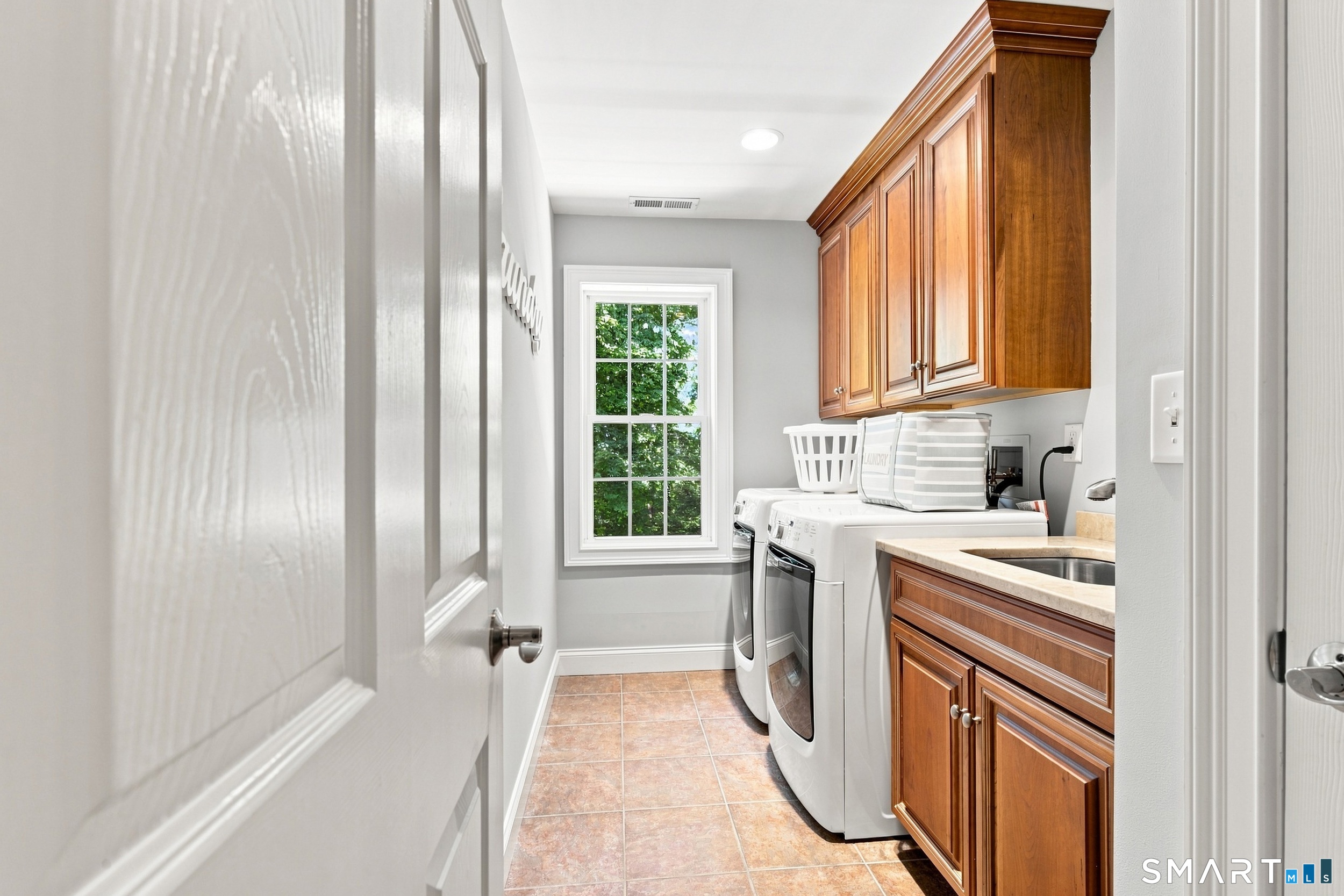 Laundry room with front-loading appliances, warm wood cabinetry, a sink, and a leafy view through the window.