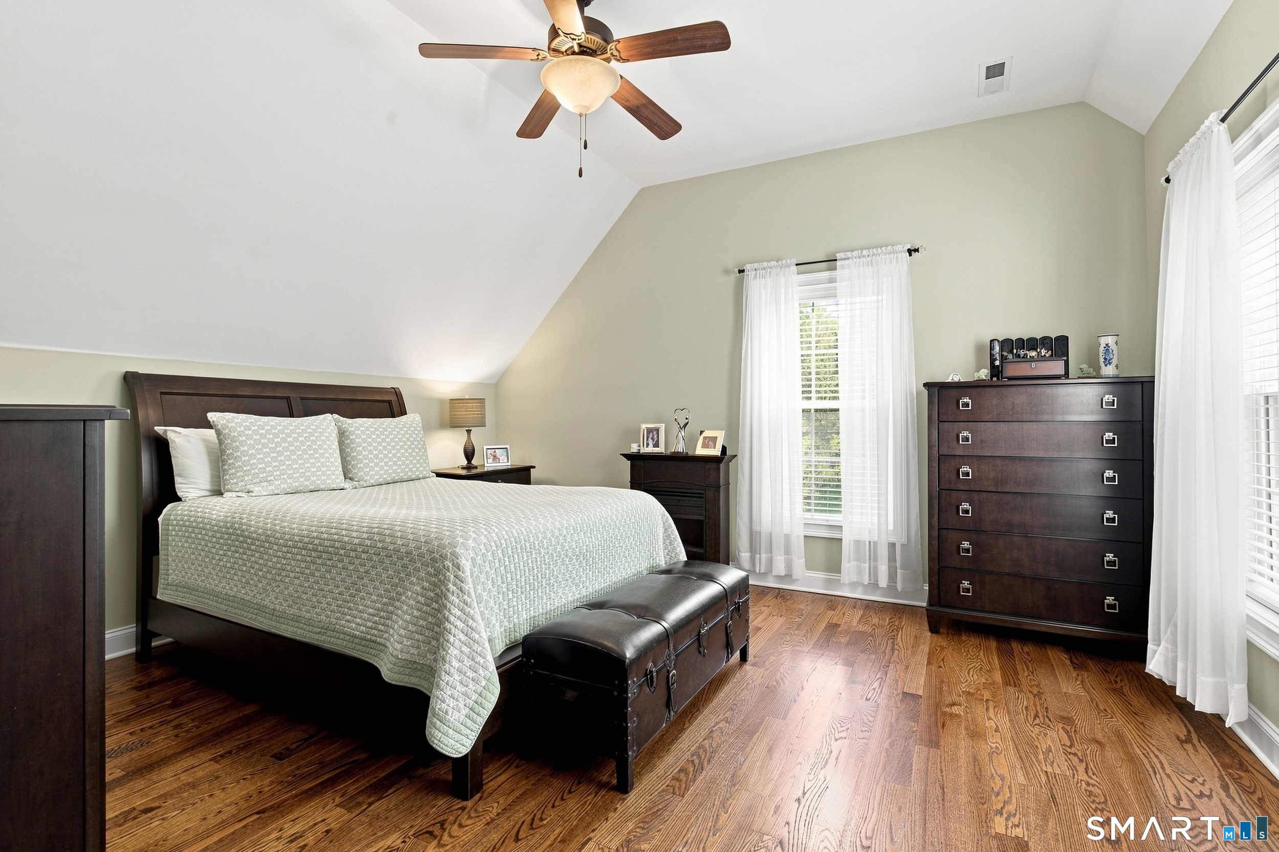 Bedroom with pale patterned bedding, dark wood furniture, hardwood floors, and tall curtained windows.