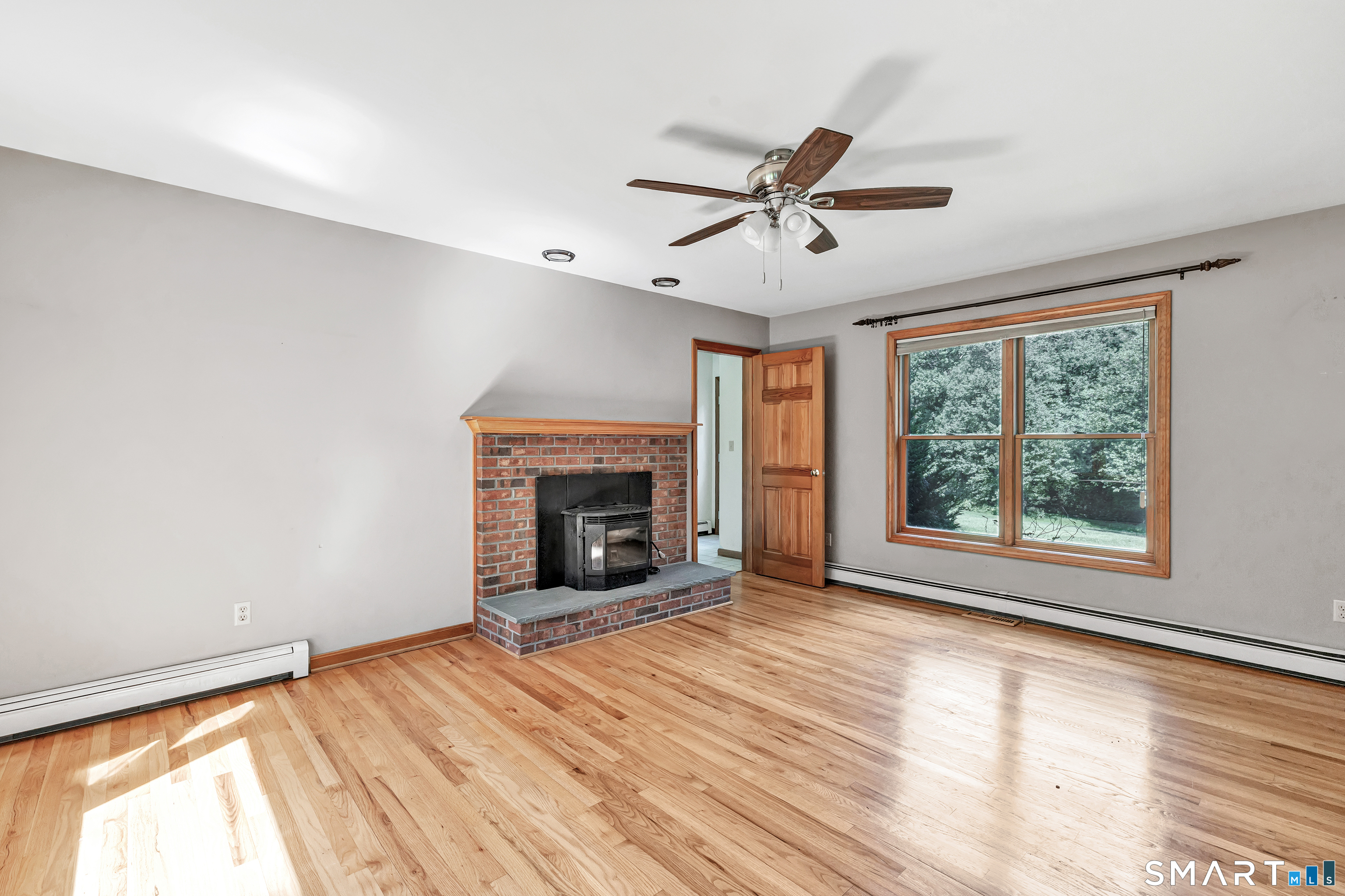 Vacant living room with hardwood floors, a brick fireplace and a large window overlooking trees.
