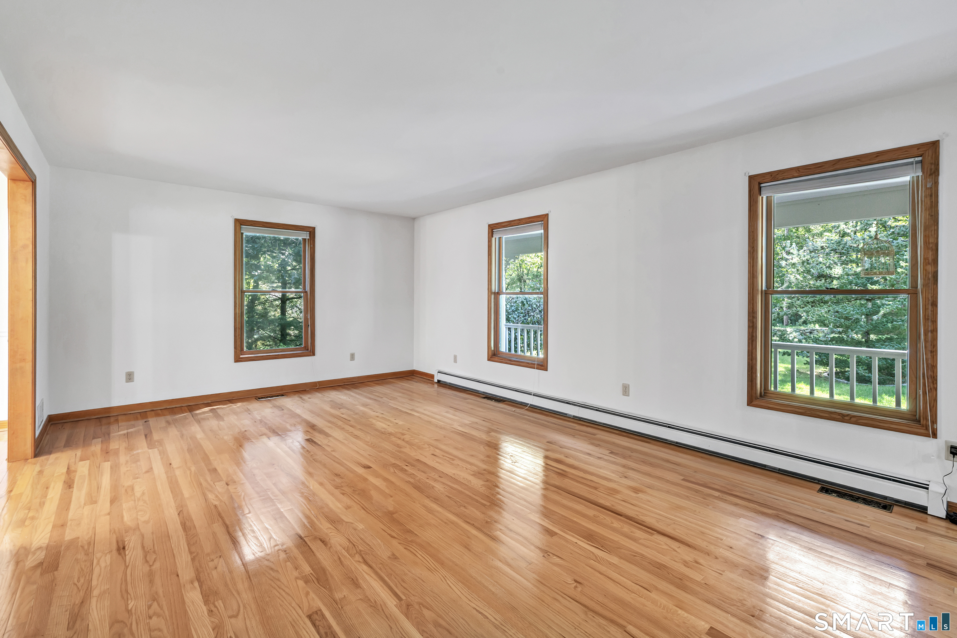 Empty living room with polished hardwood floors and three wood-trimmed windows overlooking trees.