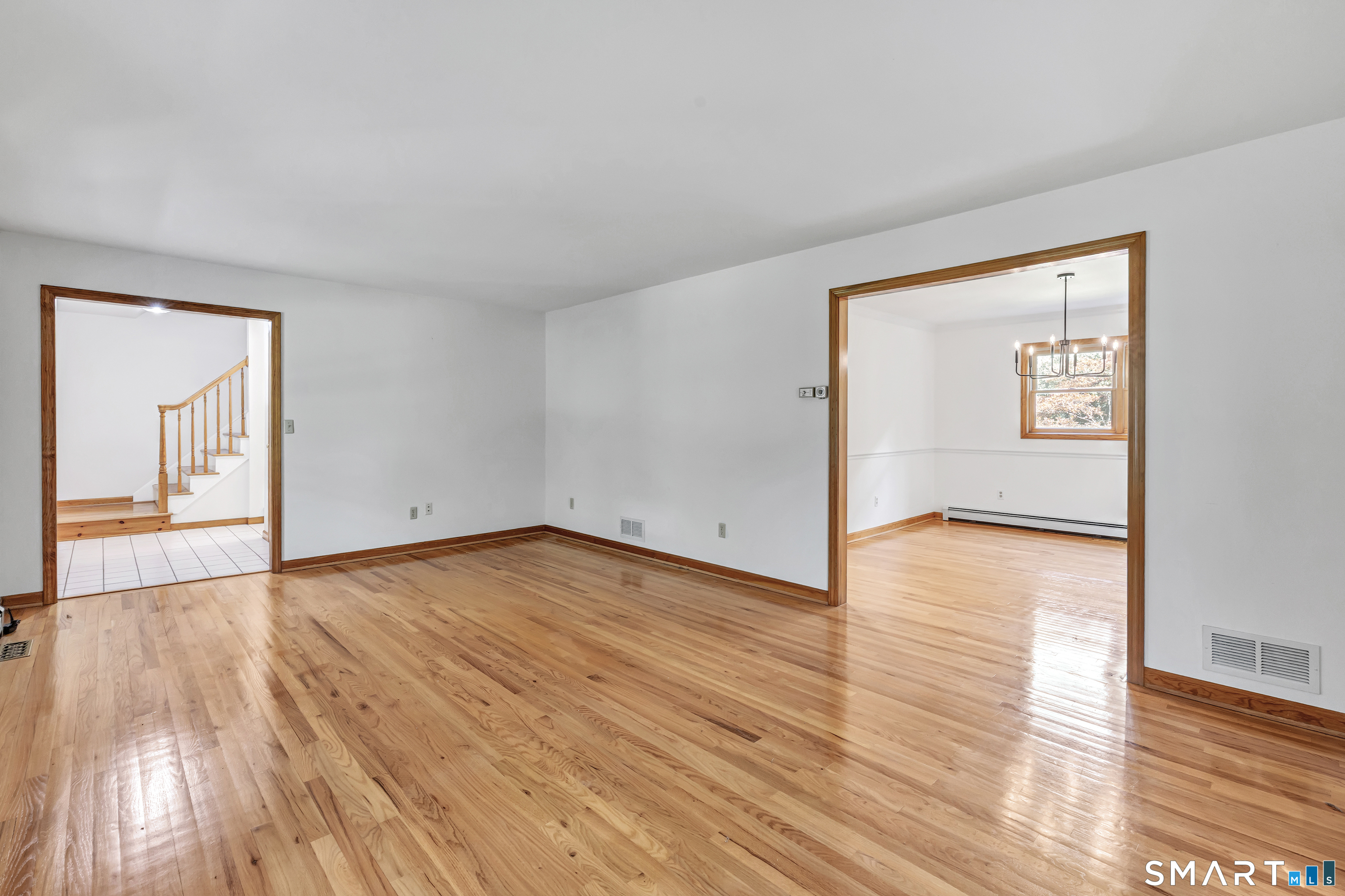 Vacant living room with polished hardwood floors, a staircase through one doorway, and a chandelier in the adjoining room.