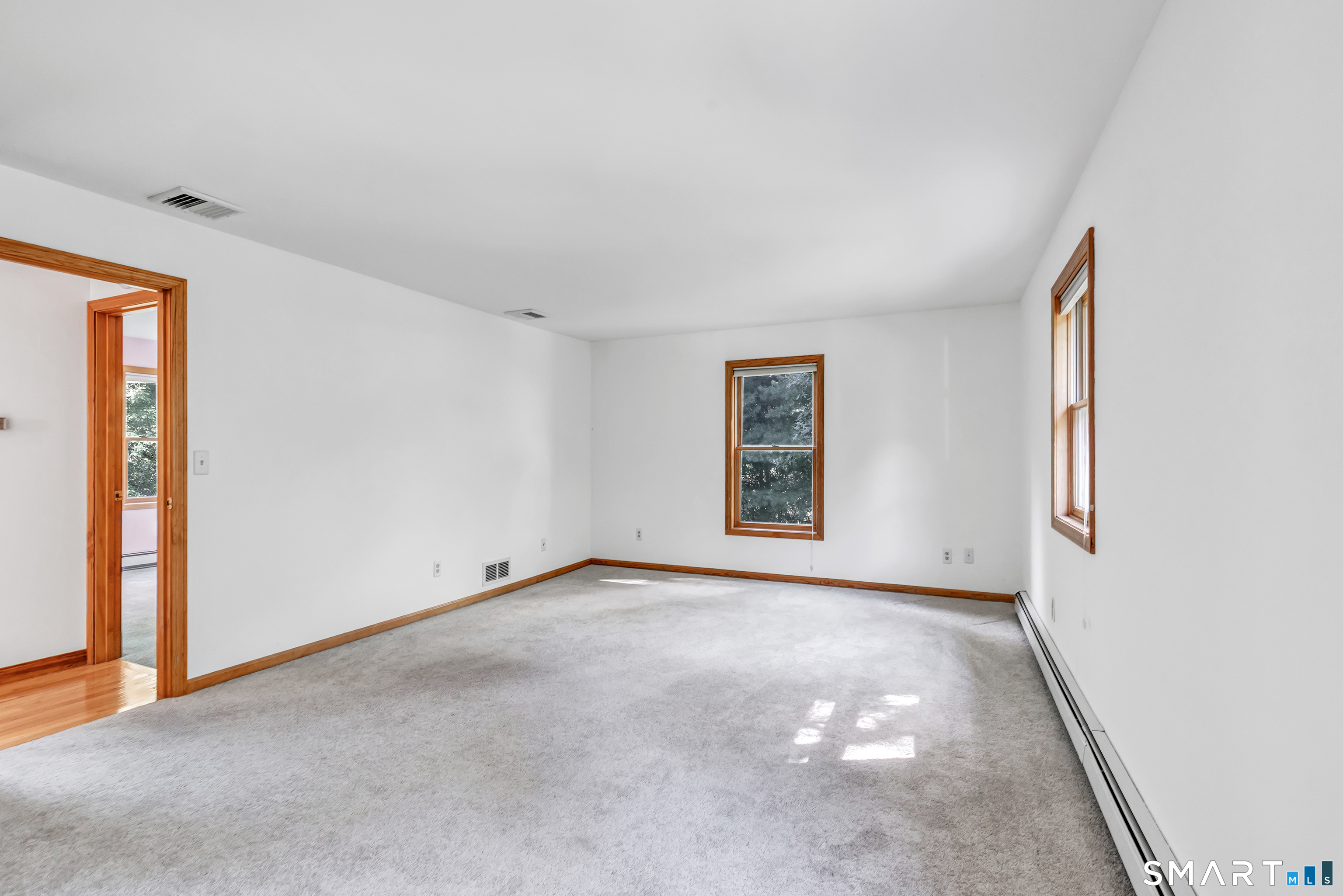 Empty carpeted room with two wood-trimmed windows and a doorway to a hardwood-floored area.