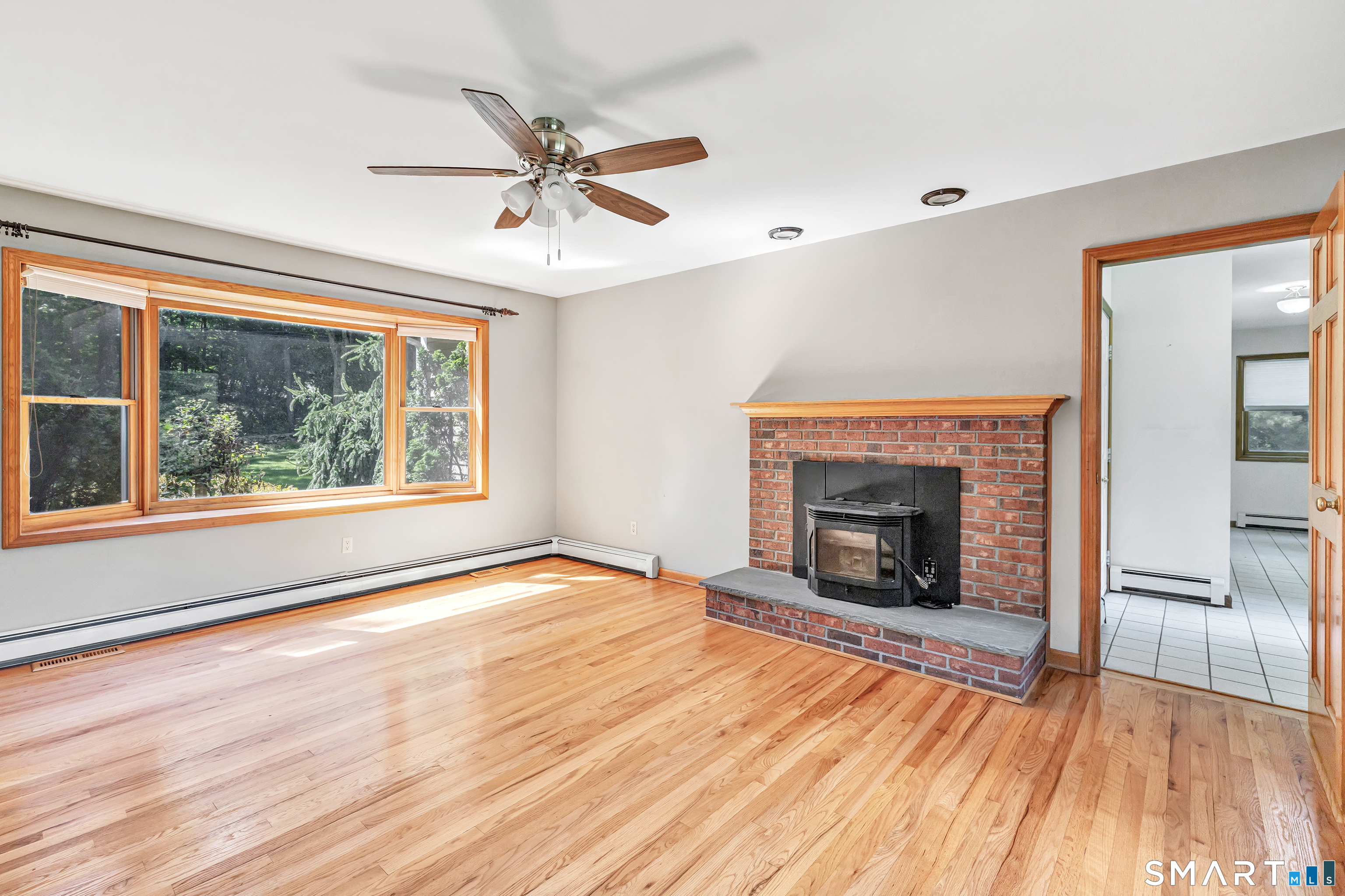 Vacant living room with hardwood floors, brick fireplace and wood stove, and a large window overlooking greenery.
