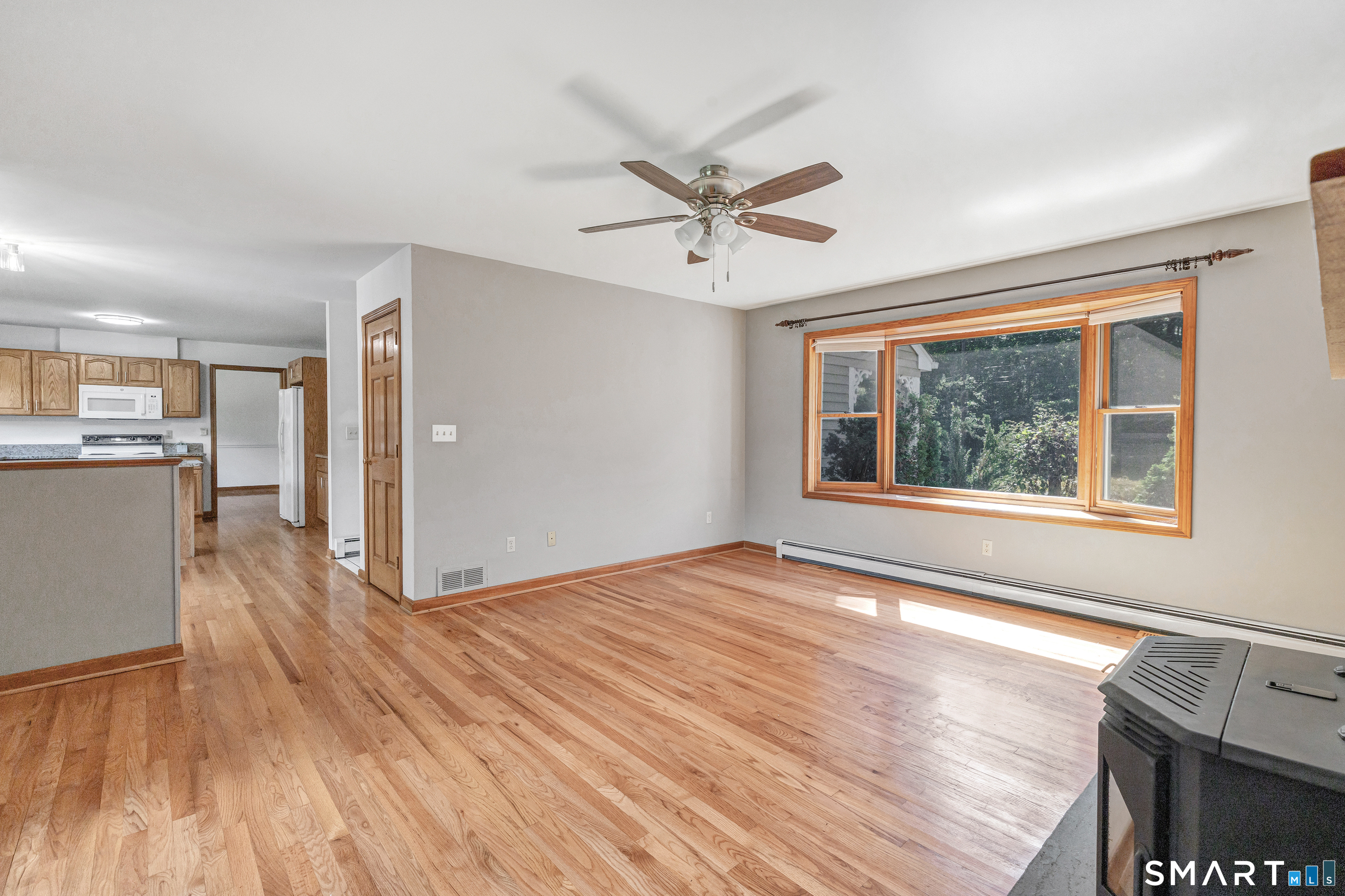 Living room with hardwood floors, ceiling fan, large window, and adjoining kitchen.